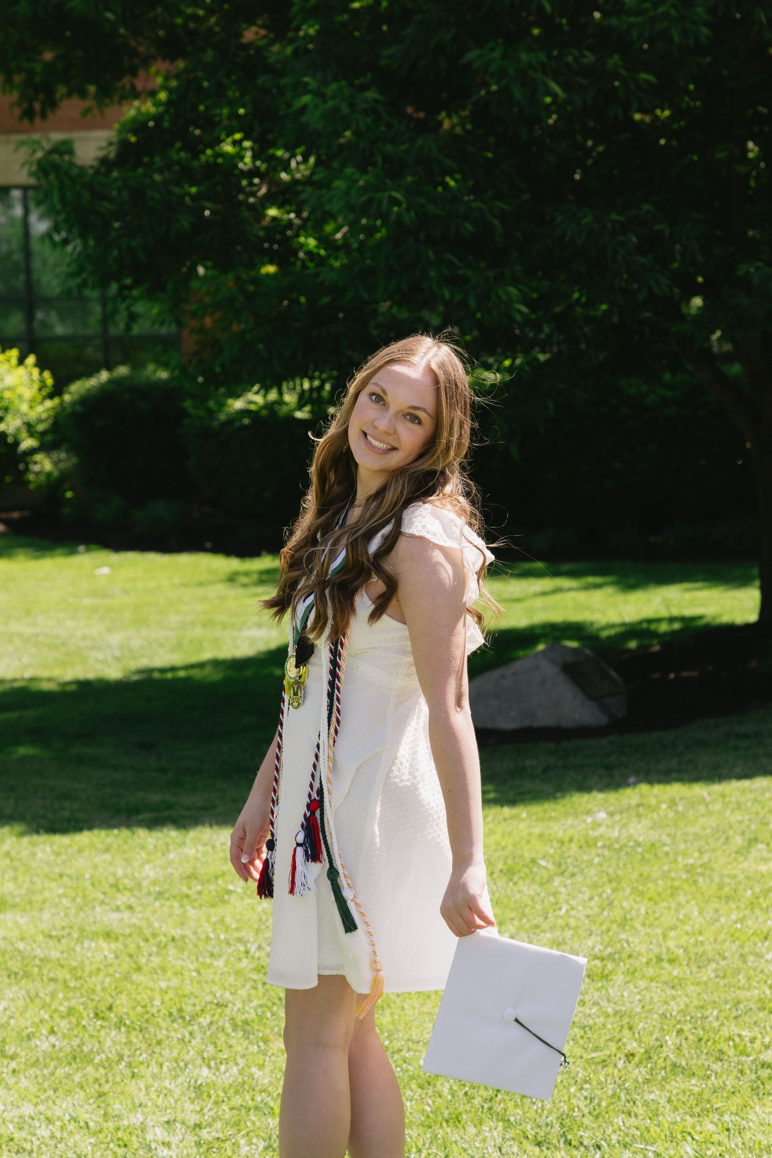 Smiling young woman in white dress holding graduation cap on green lawn with colorful honor cords.