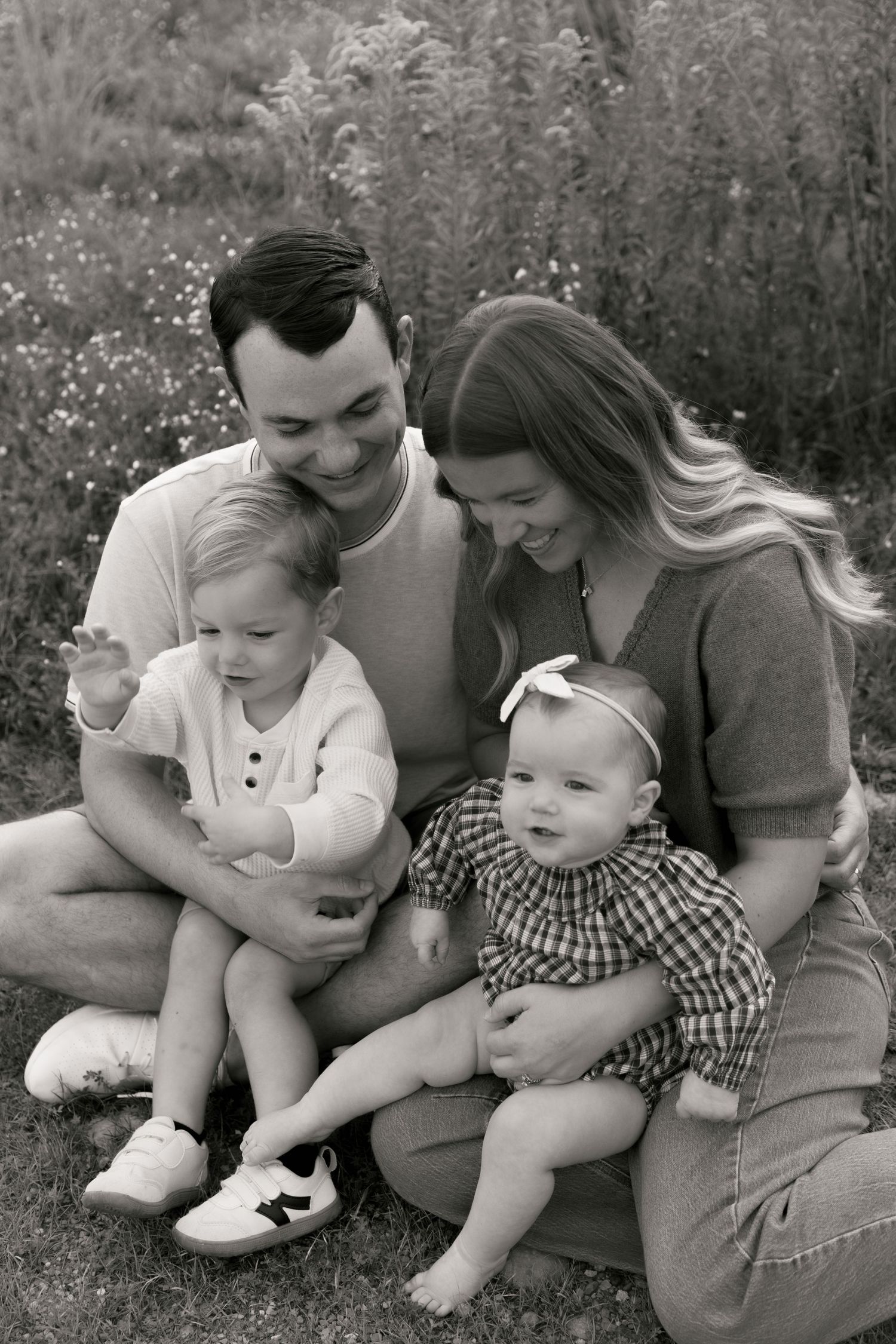 Black and white family portrait of parents sitting on grass with toddler and baby in floral field.