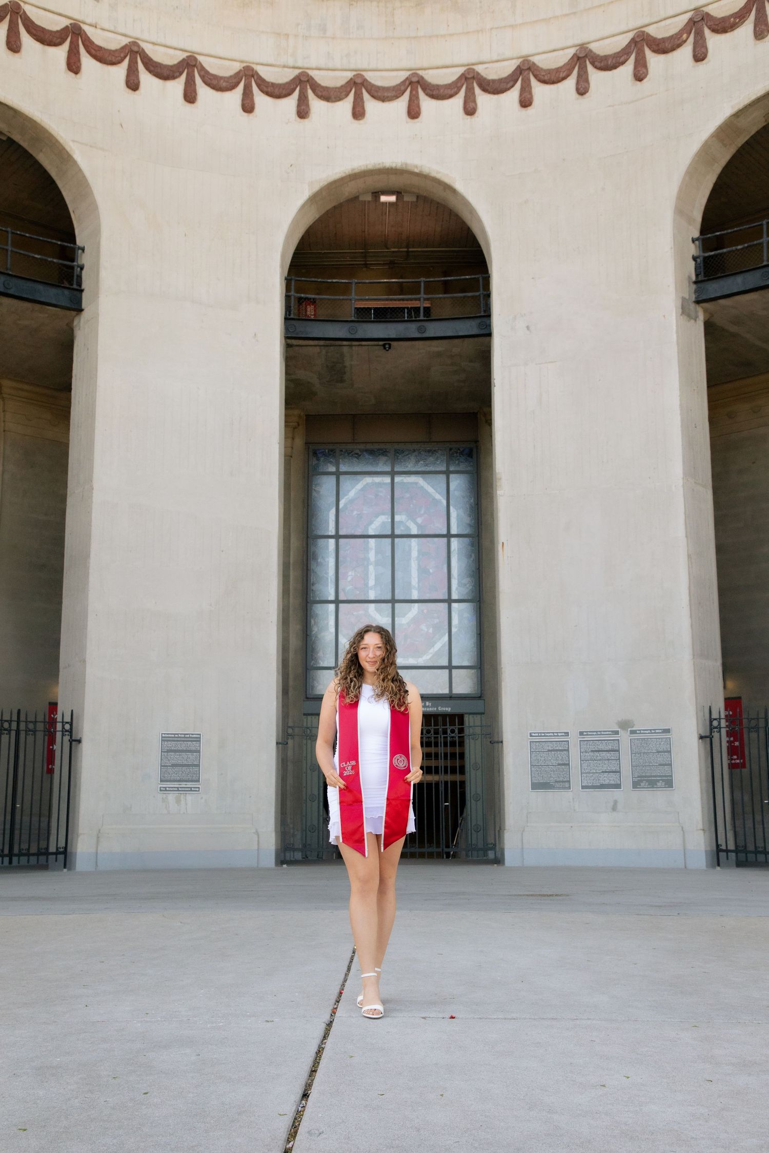 Young woman in white dress with red graduation stole poses proudly in front of a grand arched stadium entrance.