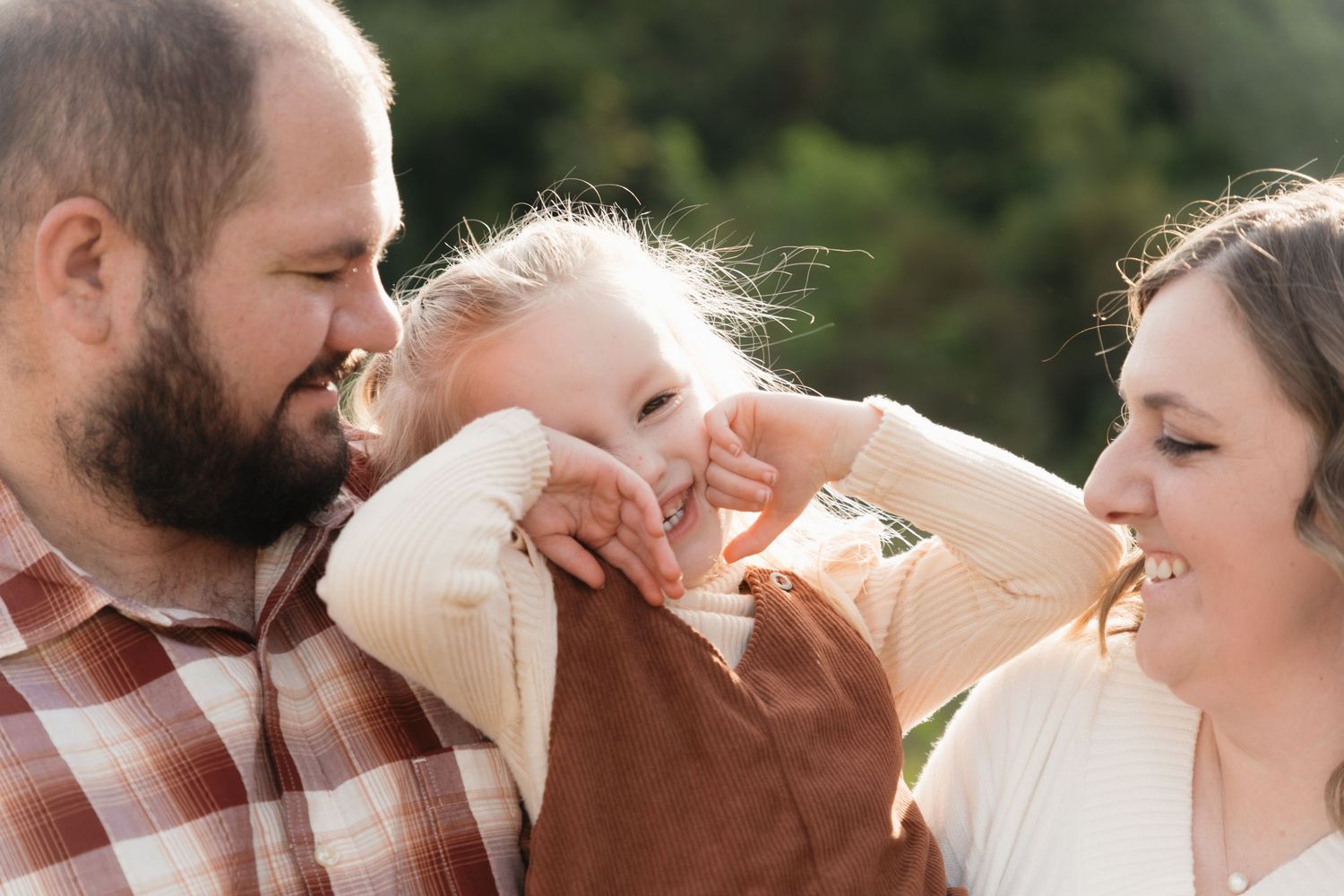 A happy young family with a toddler girl laughing outdoors in warm natural light during a family portrait session.
