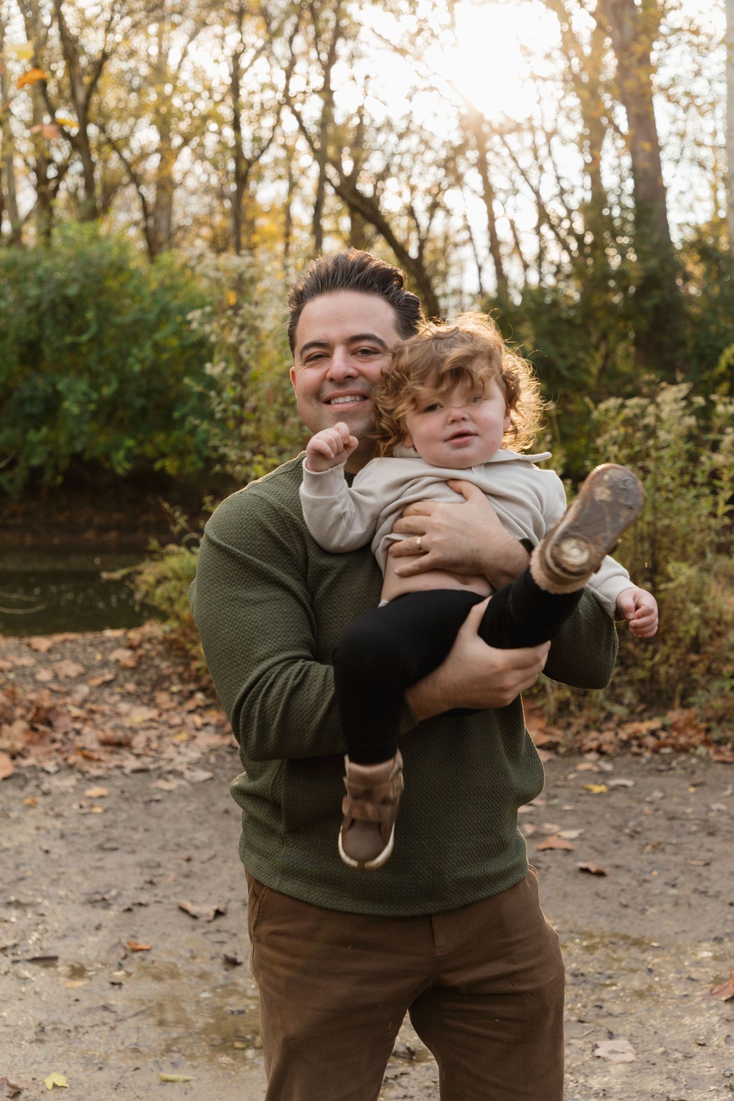 Smiling man in green hoodie holding a young toddler with curly red hair in an autumn park setting.