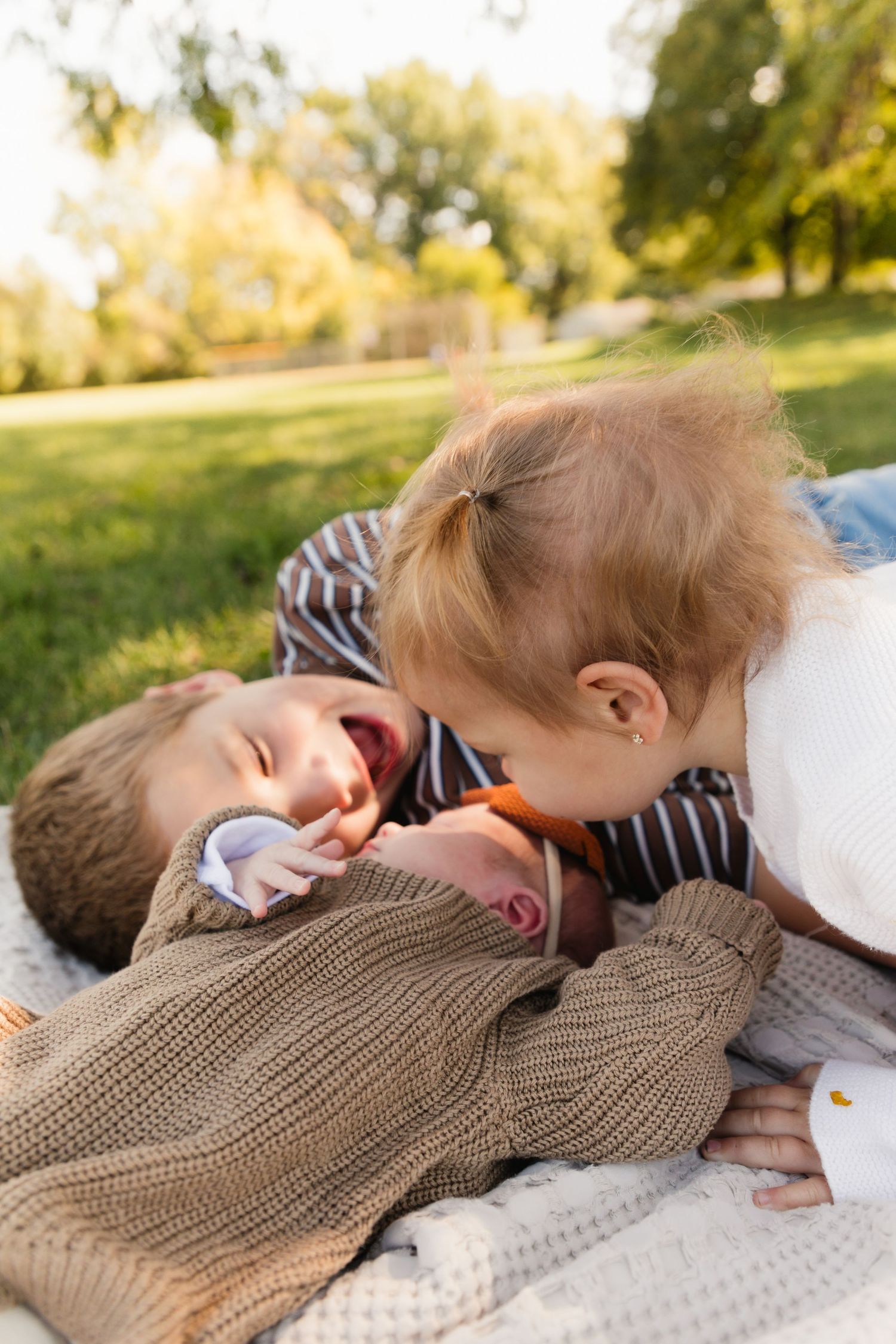 Two toddlers playing together on a blanket in a sunny park, one laughing joyfully as the other leans in close.