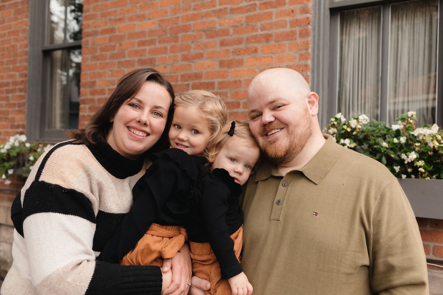 Happy family of three posing outdoors, smiling couple holding toddler in front of brick building with flowers.