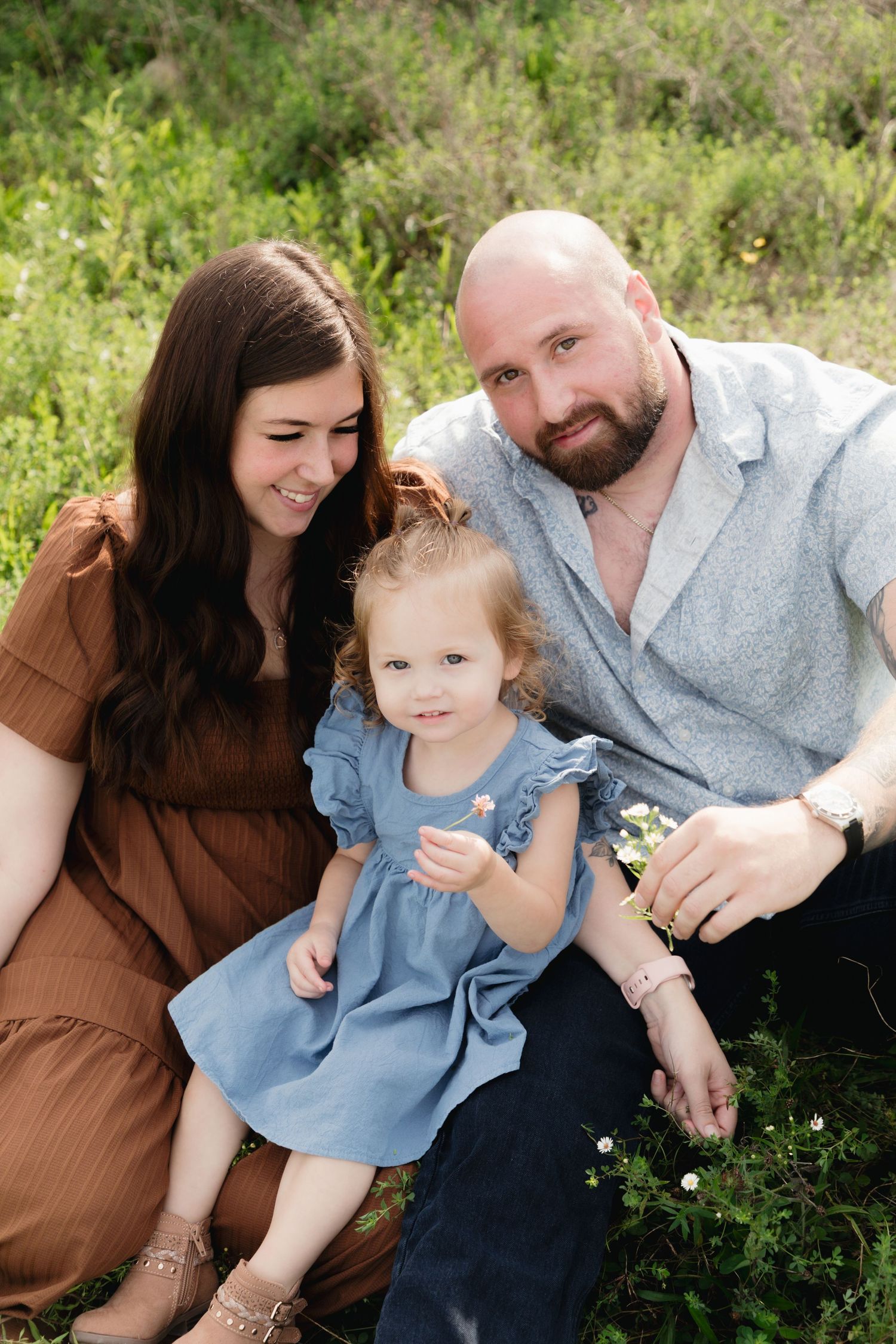 A young family with a toddler girl in a blue dress sits together in a lush green meadow during a outdoor photo session.