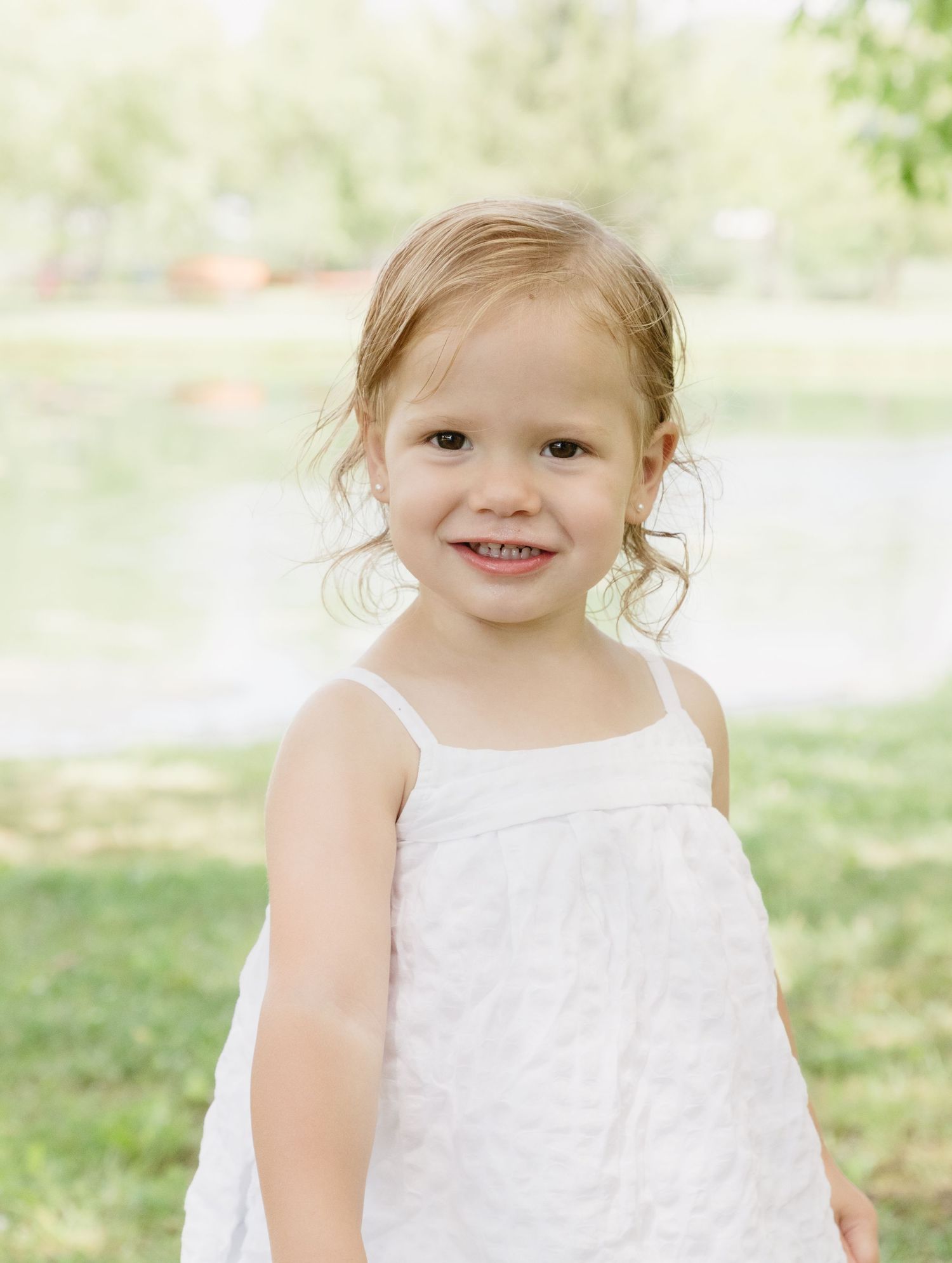 A smiling toddler girl in a white sundress stands outdoors near a pond on a sunny day.