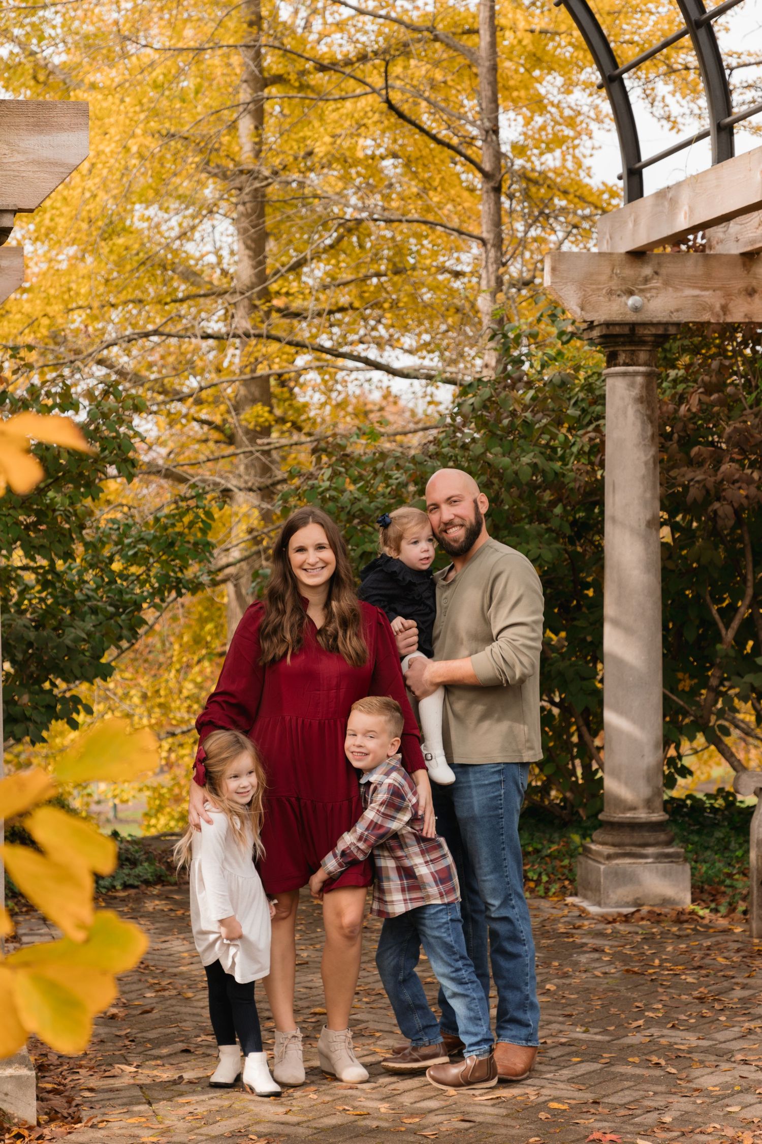 Family of five posing outdoors among vibrant fall foliage near a stone pergola during an autumn photo session.