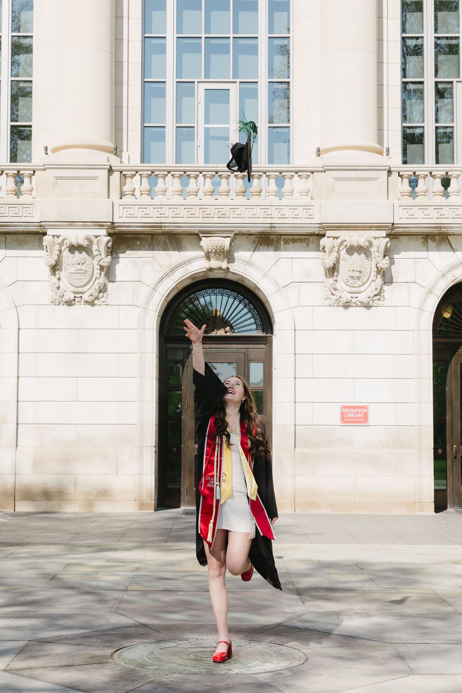 A graduate in black cap and gown with red sash tosses her cap in front of a grand university building.