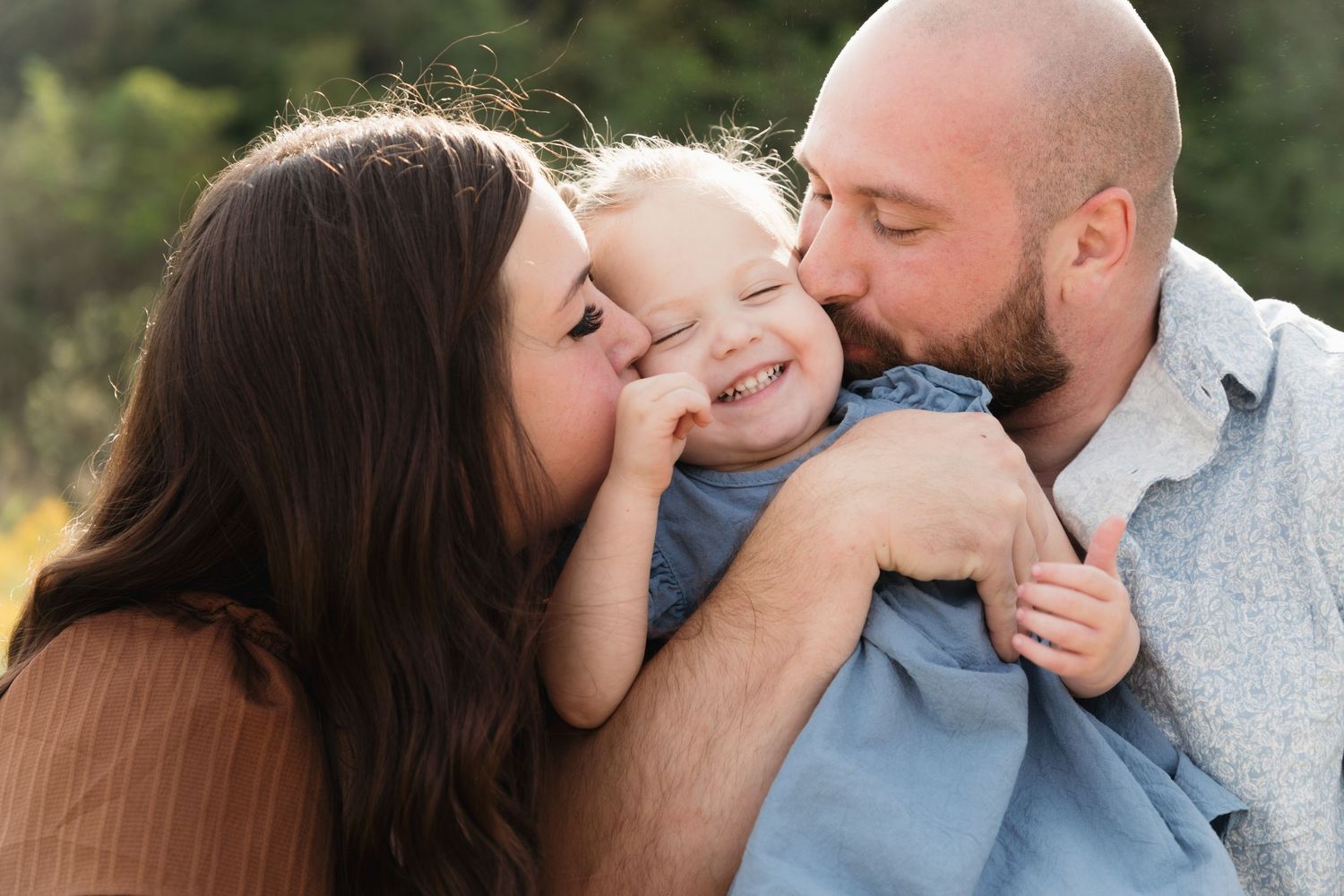 Parents kissing their smiling toddler on the cheeks outdoors in a warm, candid family portrait.