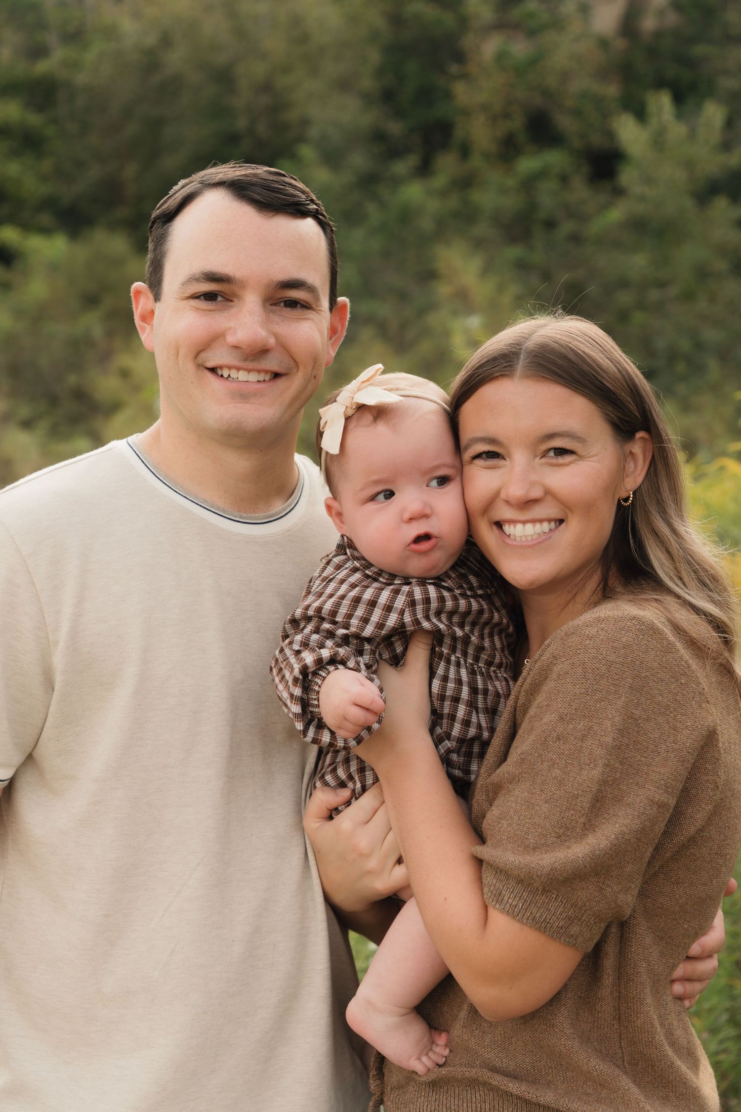 A smiling young family with a baby girl in a plaid outfit poses outdoors with green trees in the background.