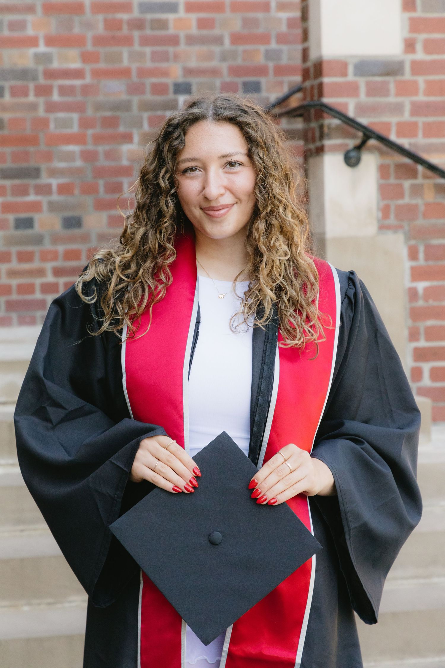 Young woman in black graduation gown with red sash holds cap, smiling in front of brick building.
