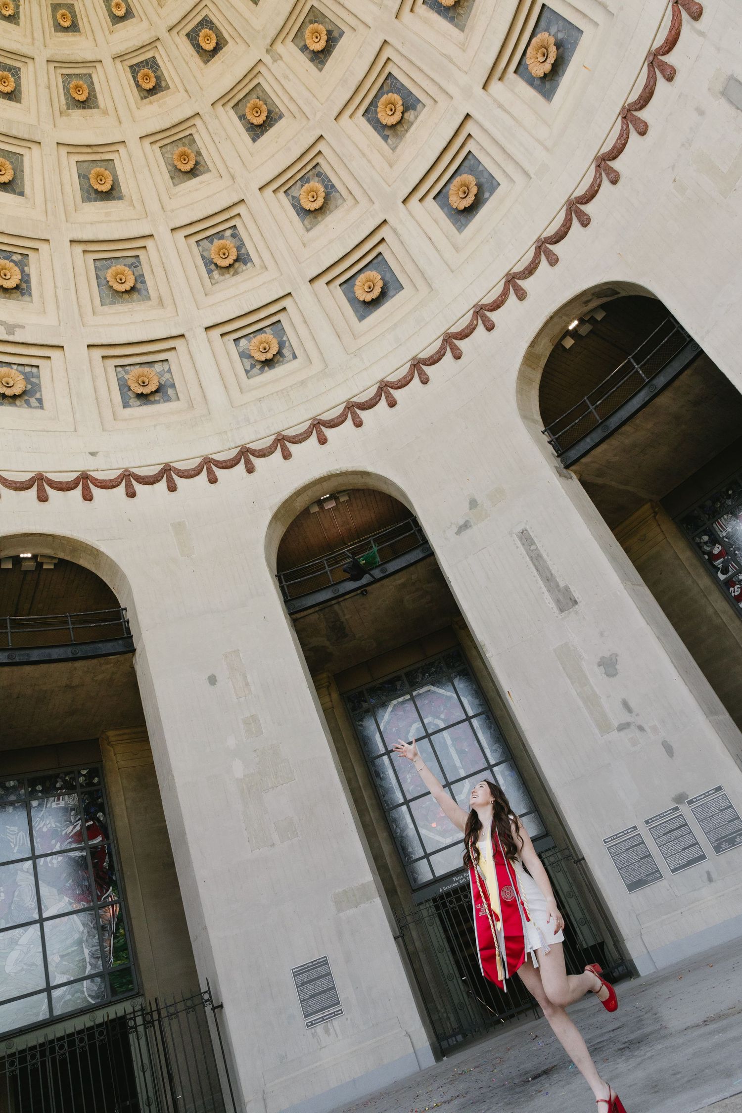 Graduate in red and white dress poses beneath ornate domed ceiling with arched windows of a grand classical building.