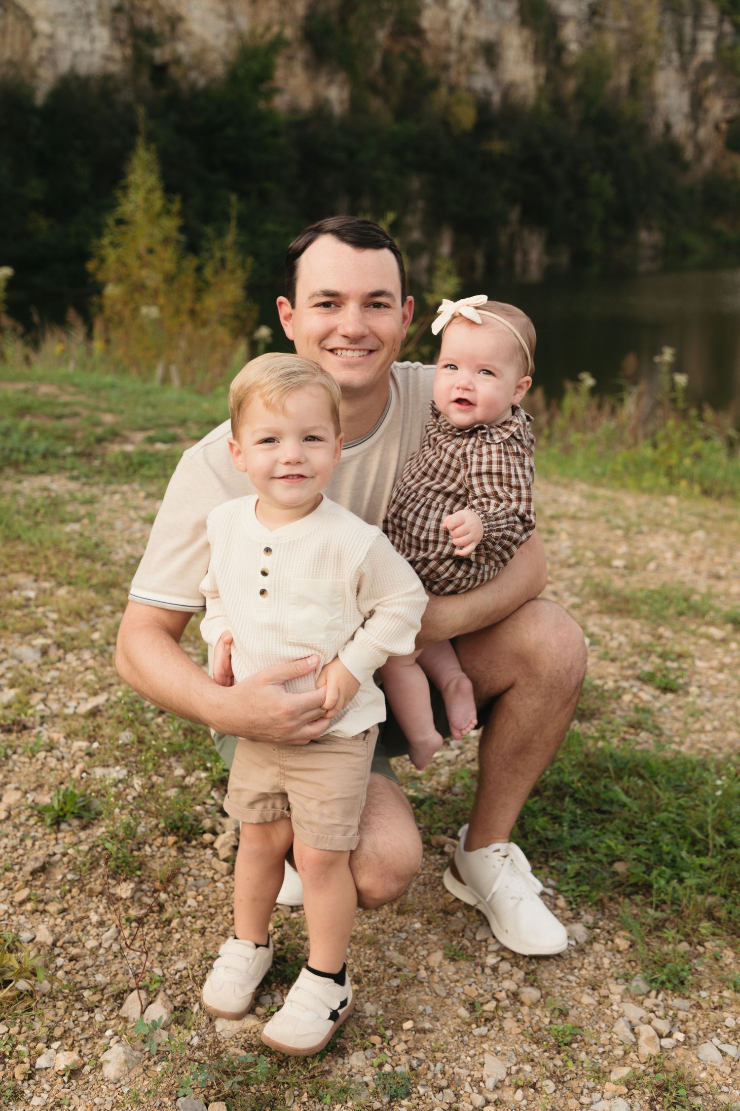 A smiling father kneels outdoors holding a baby girl and hugging a toddler boy near a scenic lake and rocky cliffs.