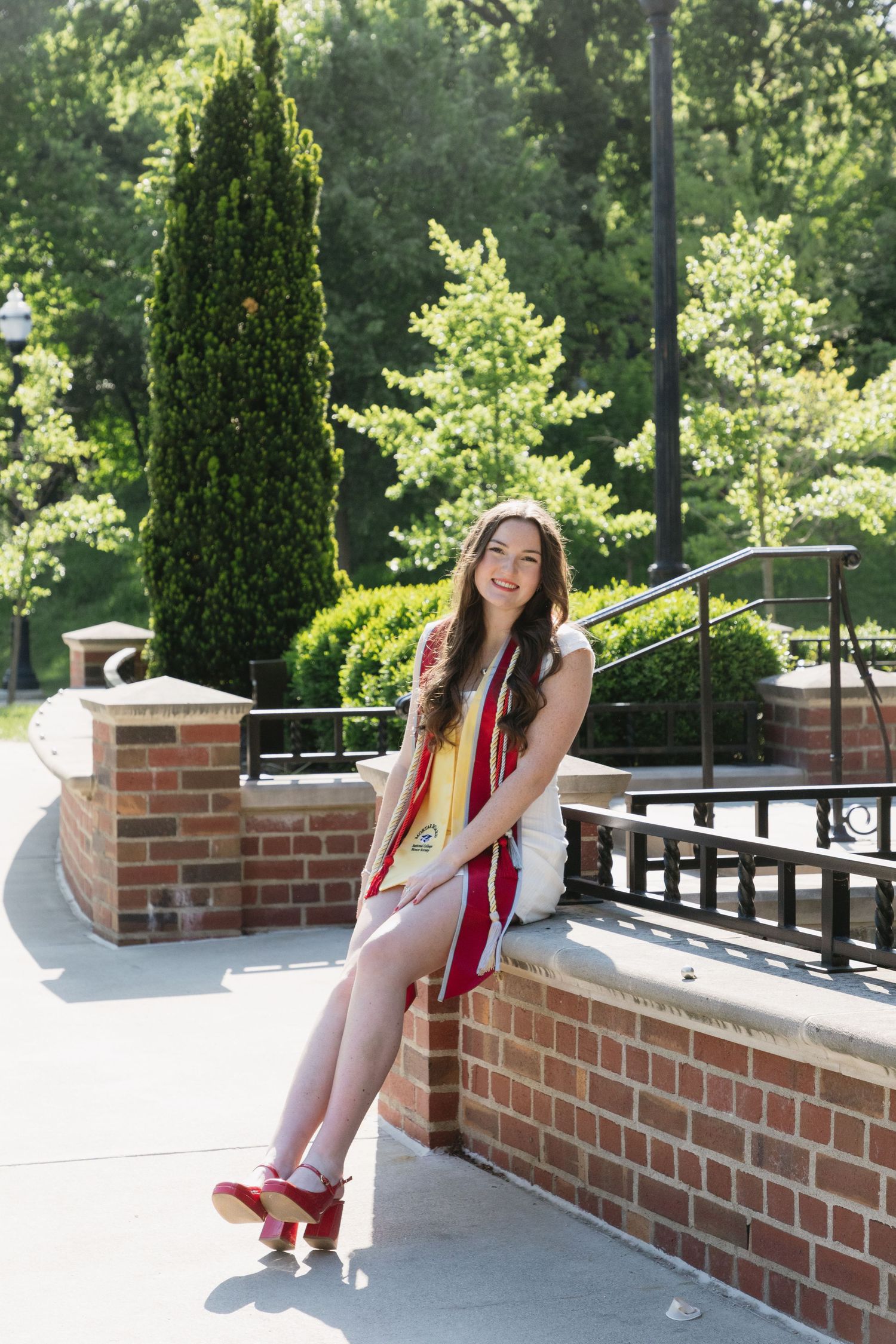 Smiling young woman in graduation attire sitting on brick wall in sunny campus outdoor setting.