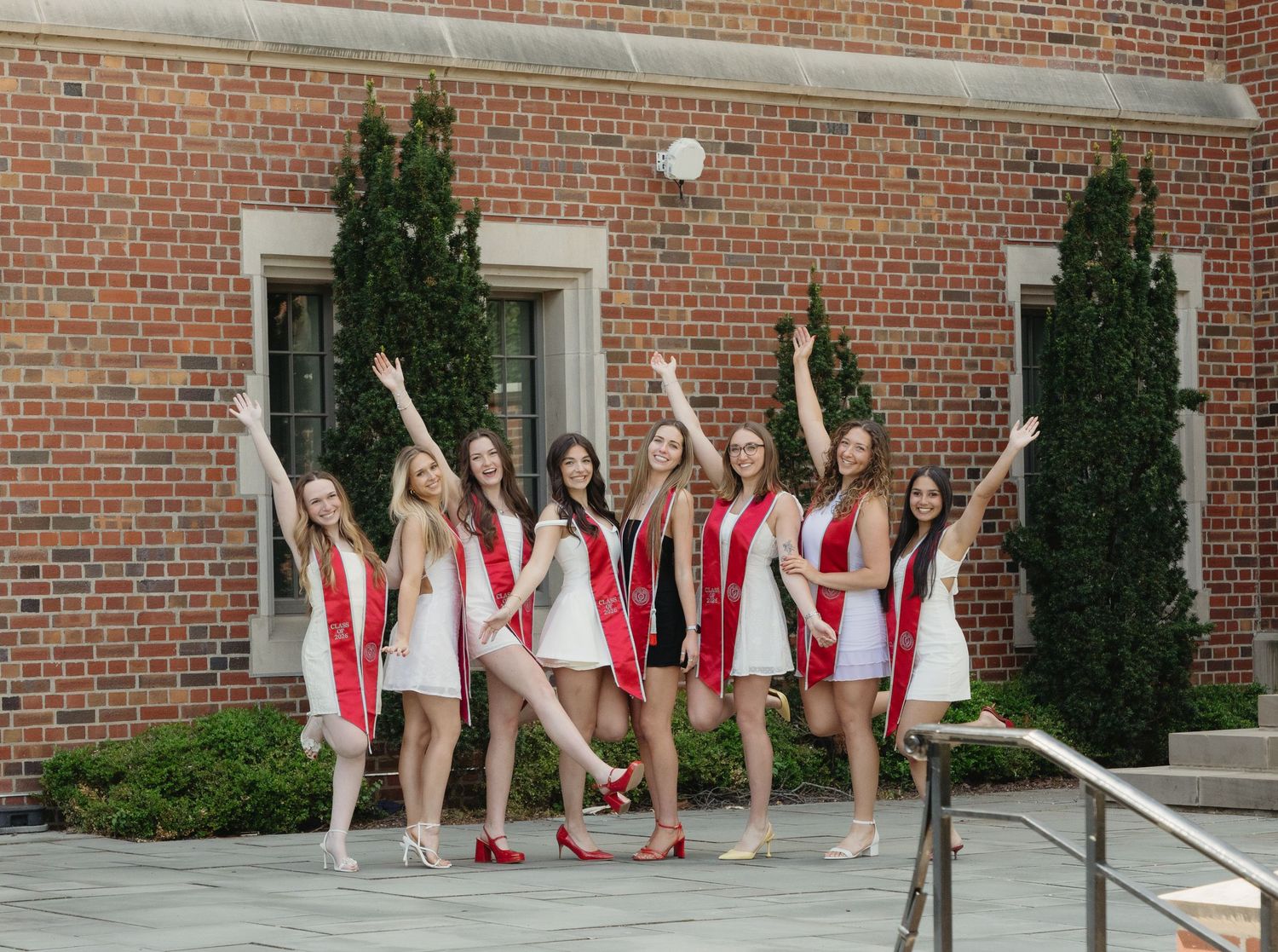 Group of college graduates in white dresses with red sashes celebrating outdoors near a brick building.