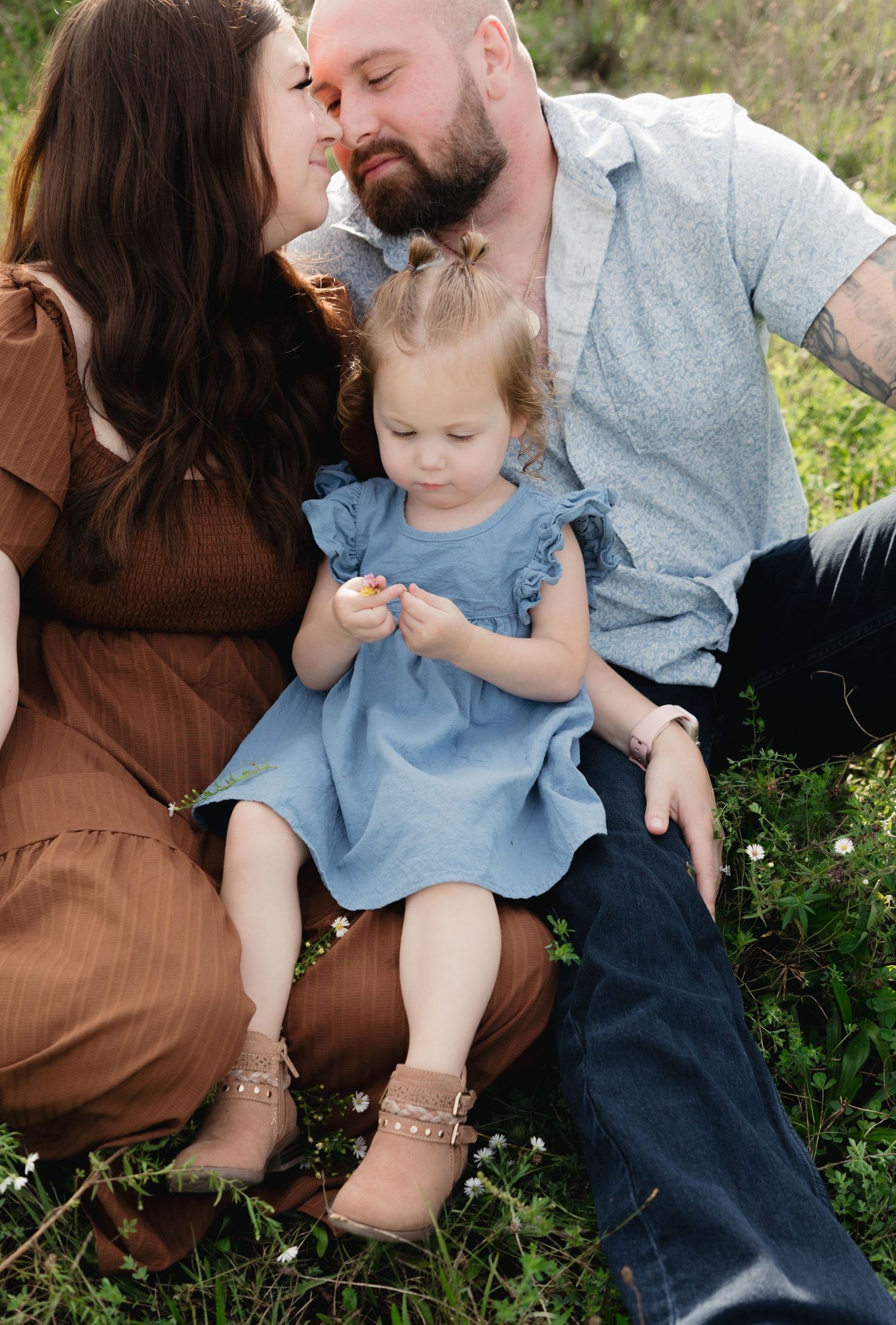 A family of three sits in a grassy field, parents kissing while toddler girl in blue dress examines a flower.