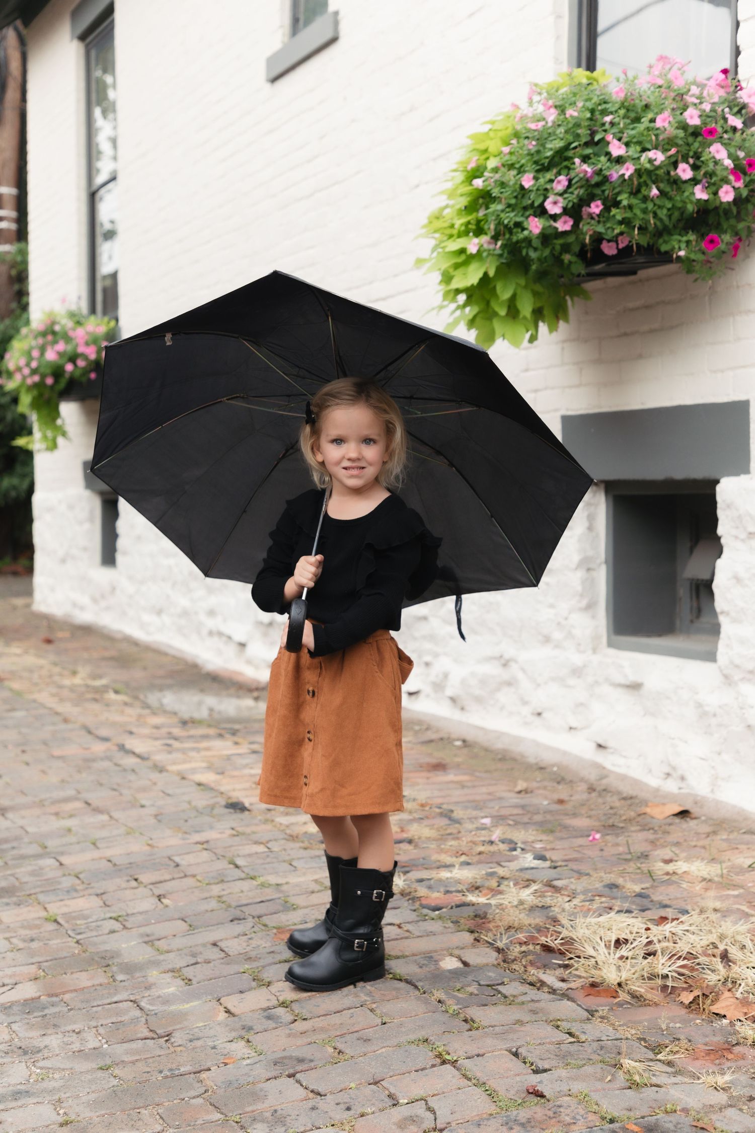 Young girl in black top, brown skirt, and boots holds a large black umbrella on a cobblestone street.
