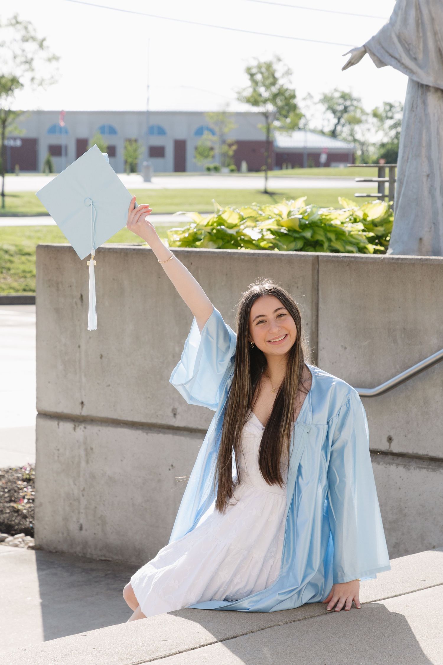 Smiling graduate in light blue cap and gown sitting outdoors, raising her graduation cap in celebration.
