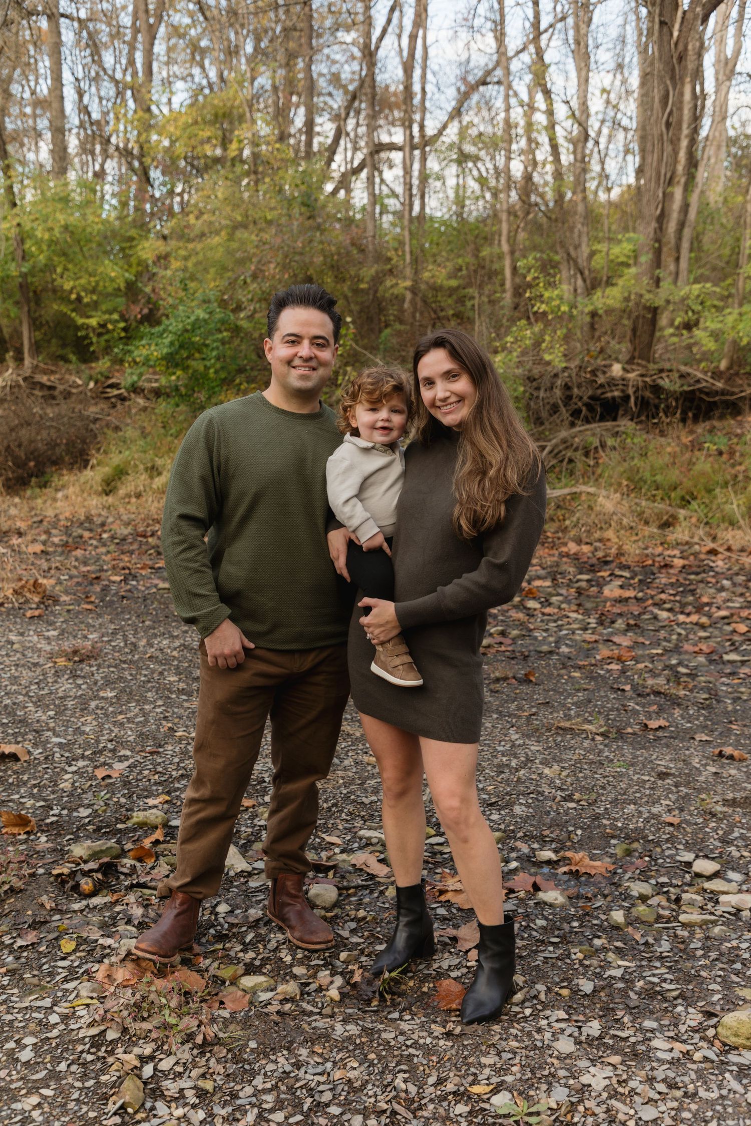 A happy young family of three poses outdoors on a rocky path surrounded by autumn trees.
