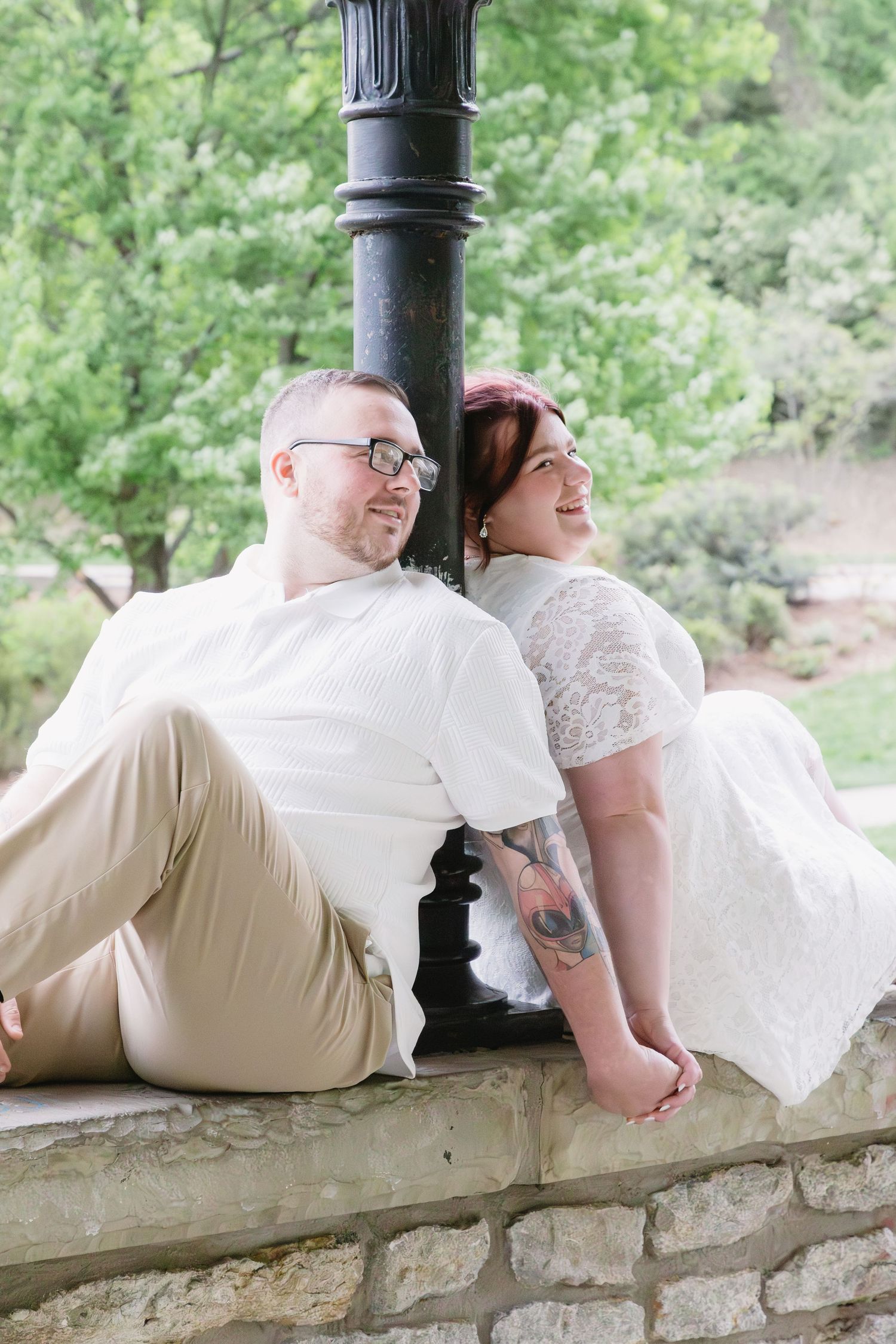 A couple in wedding attire sits back-to-back against a lamppost on a stone wall, surrounded by lush greenery.