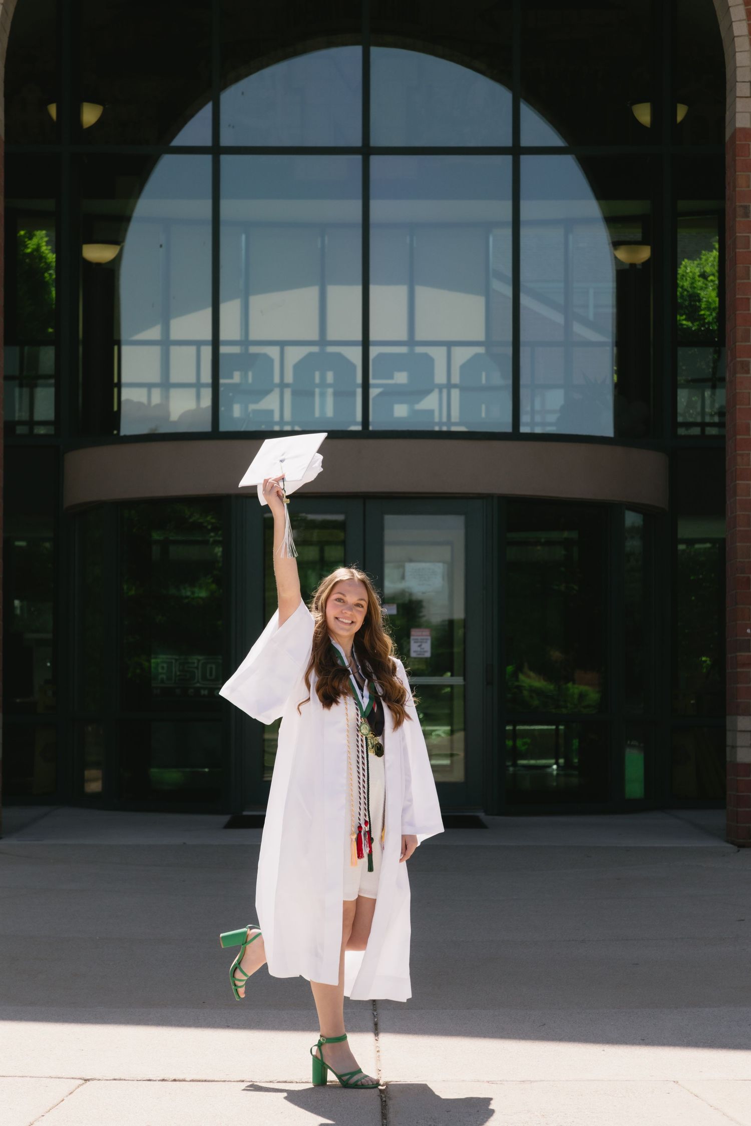 Smiling graduate in white cap and gown raises her diploma in front of a modern school building entrance.