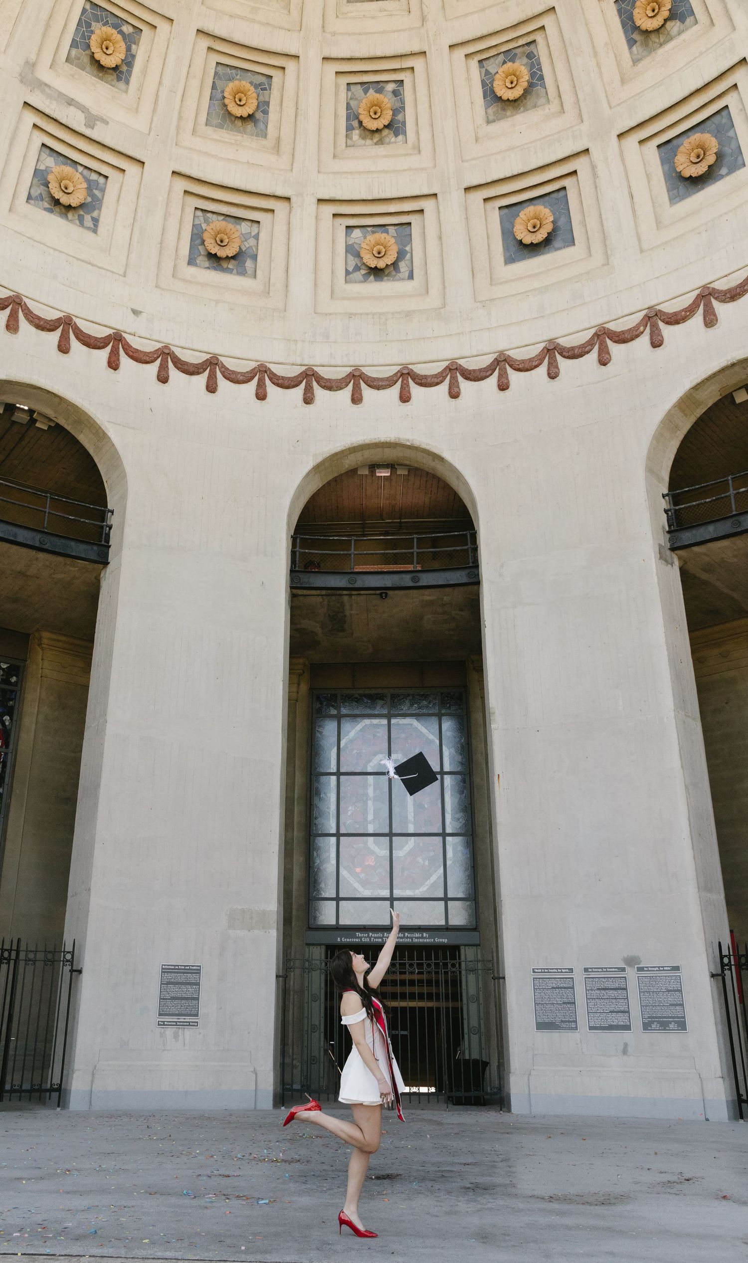Graduate in white dress and red heels tosses cap in front of ornate domed building entrance.