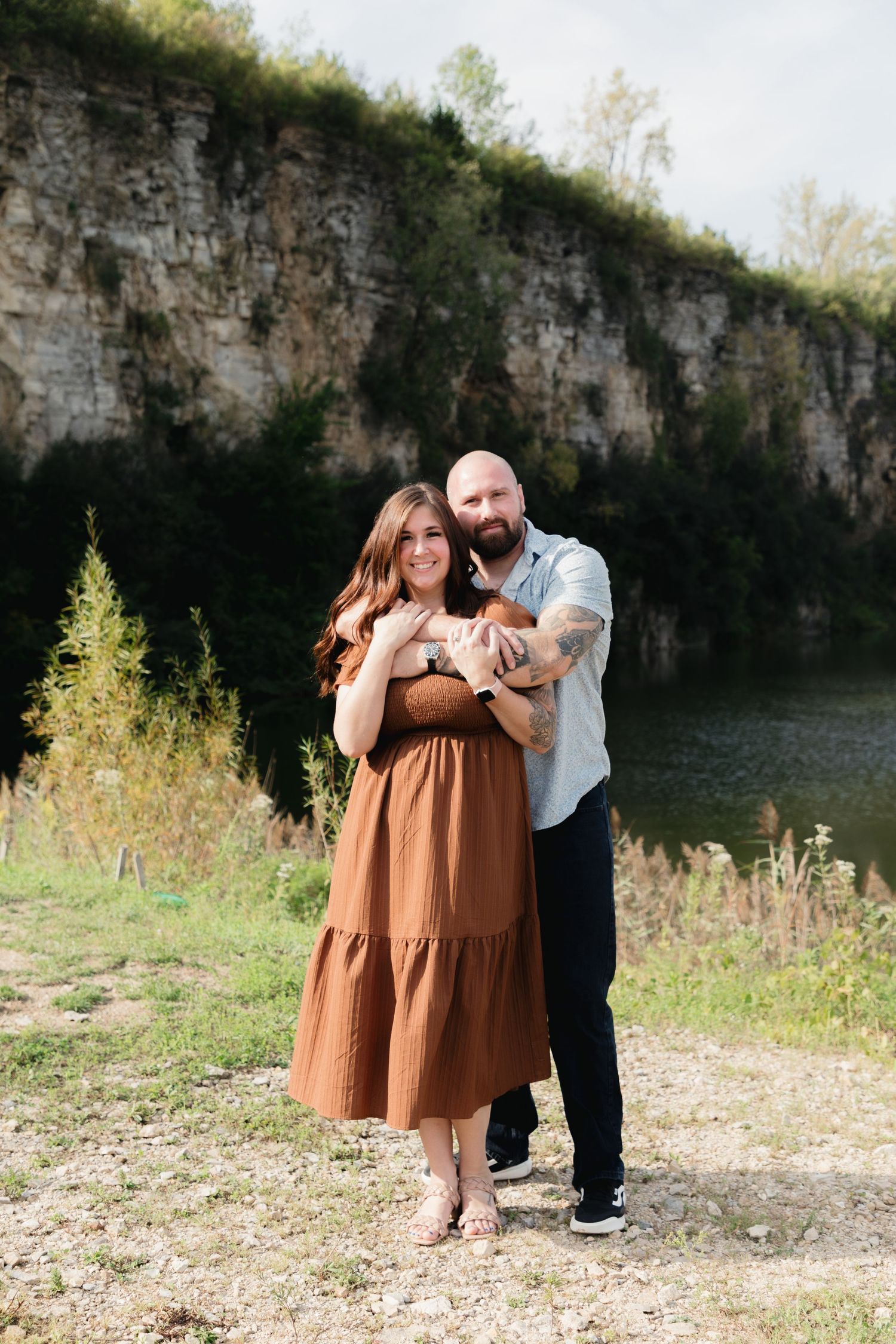 A couple embraces outdoors near a rocky cliff and lake, woman in brown dress, man in light blue shirt with tattoos.