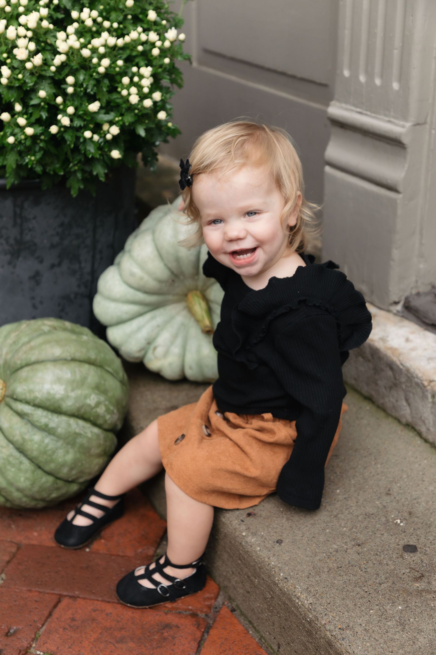 Smiling toddler girl in black top and brown skirt sitting on steps beside green pumpkins and white mums.