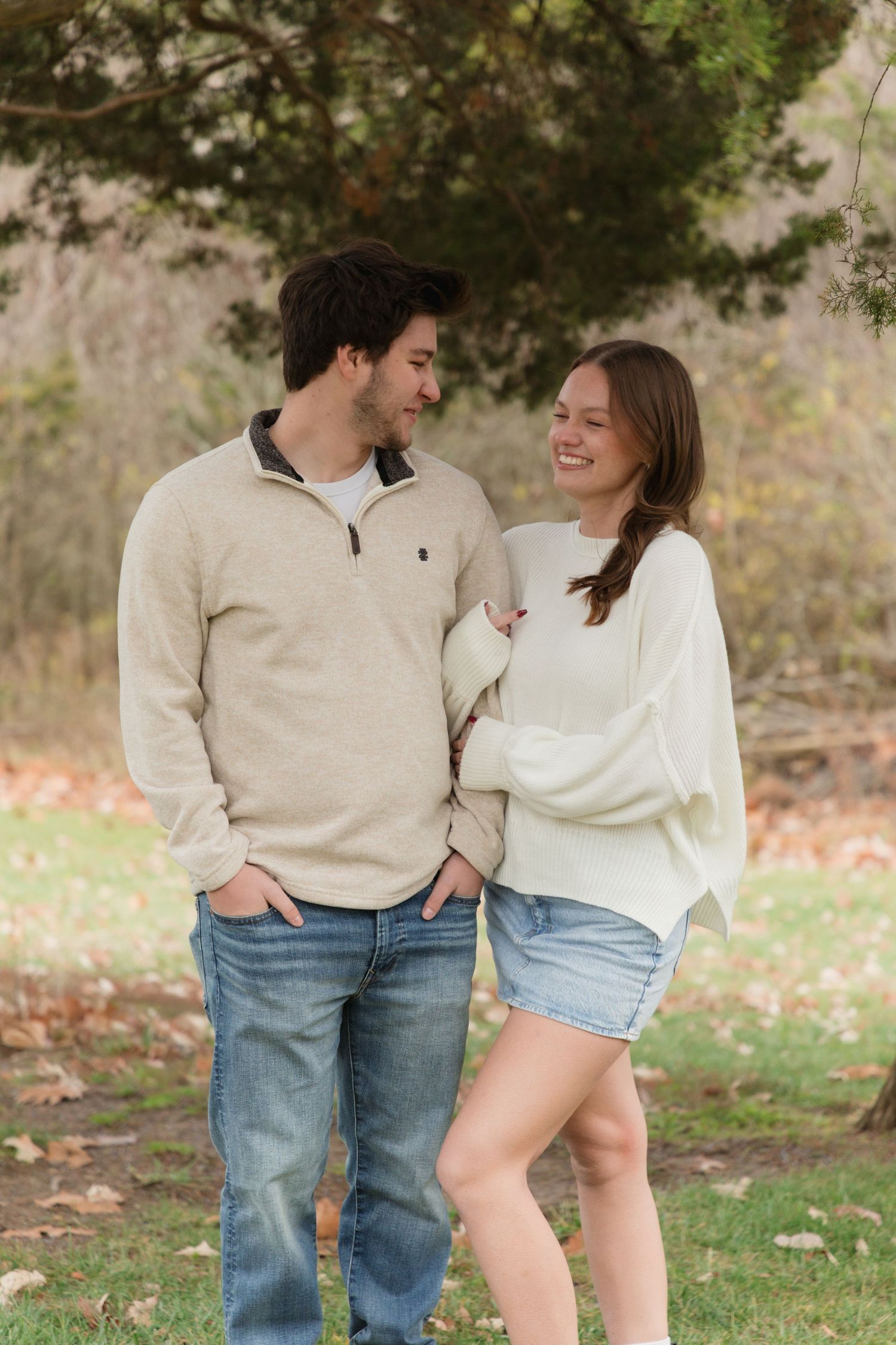 A smiling couple in casual sweaters and jeans share a warm moment outdoors in an autumn park setting.