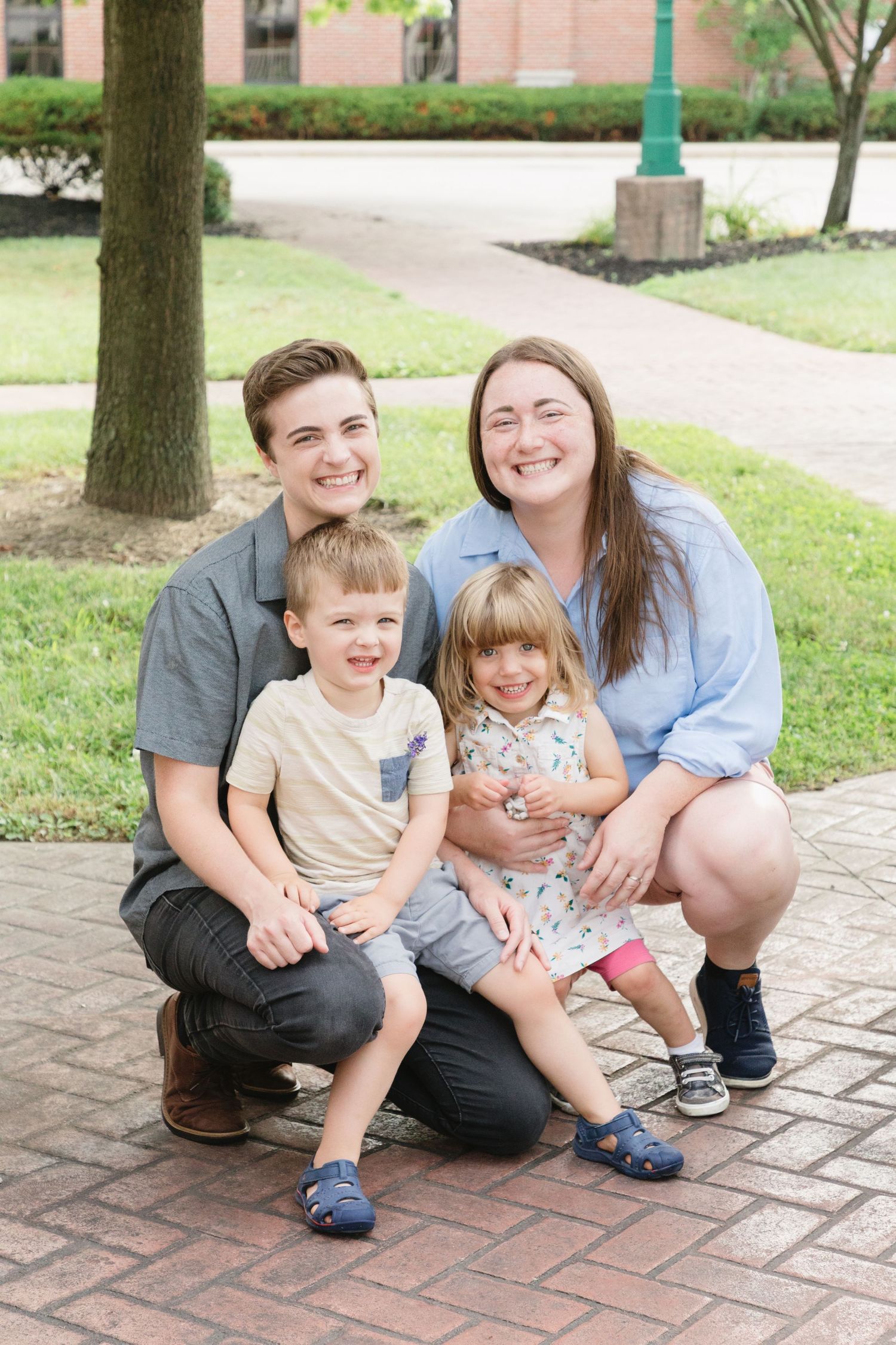 A smiling family of four poses outdoors on a brick path, with two young children seated in front of two adults.