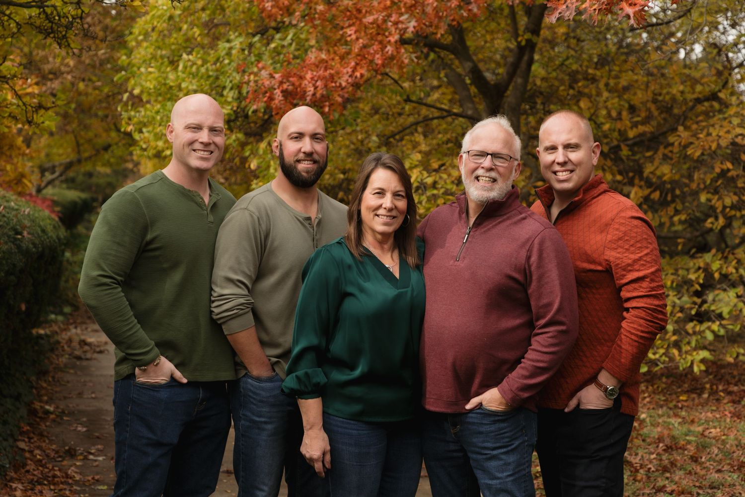 Family of five smiling outdoors among vibrant autumn foliage, dressed in coordinated fall colors for a portrait.