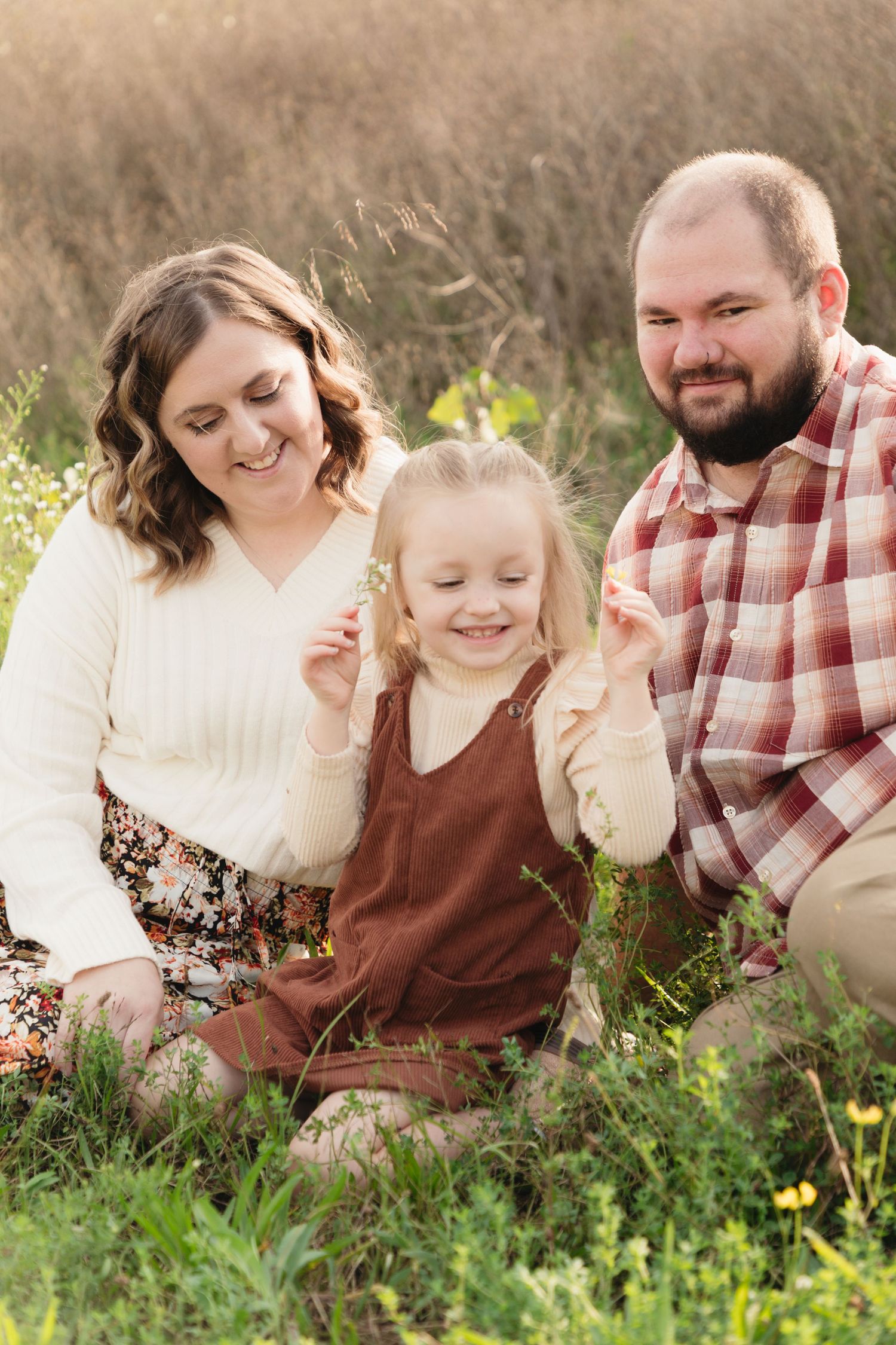 A happy family of three sits in a sunny field, parents smiling at their young blonde daughter holding wildflowers.