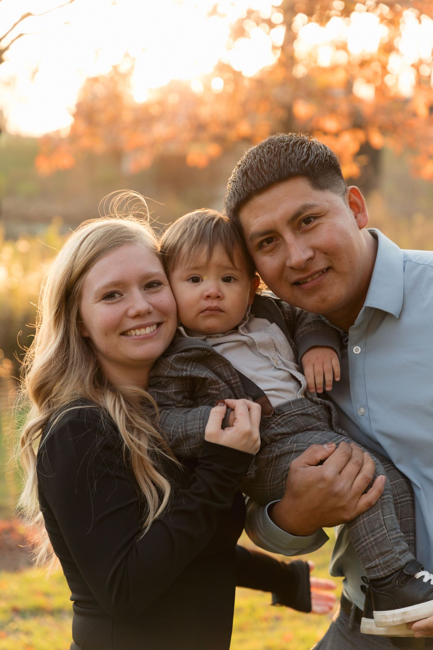 A young family with a blonde woman, a man, and their toddler posing outdoors during golden autumn sunset.