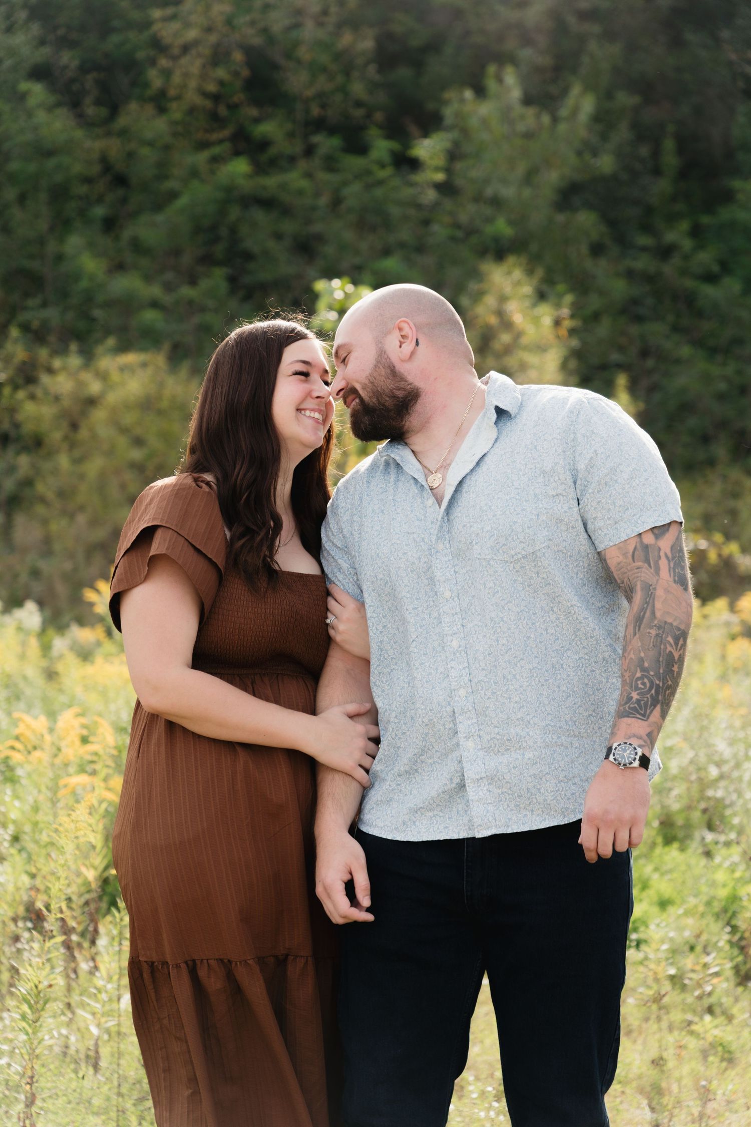 A couple shares a tender moment in a sunlit field, the man with tattoos leaning in close to the smiling woman in a brown dress.