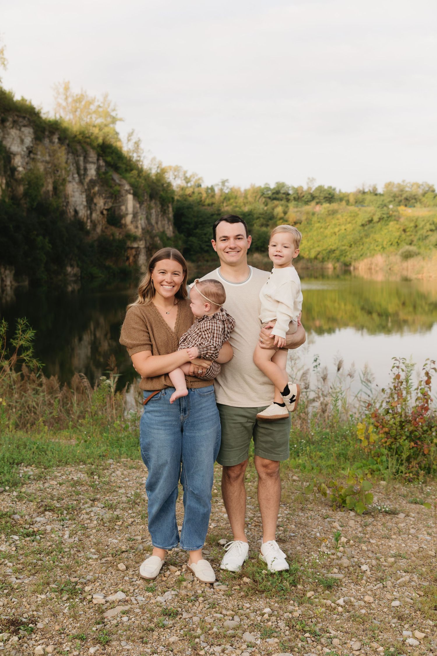 Young family of four posing outdoors near a scenic lake and rocky cliffs, parents holding two young children.