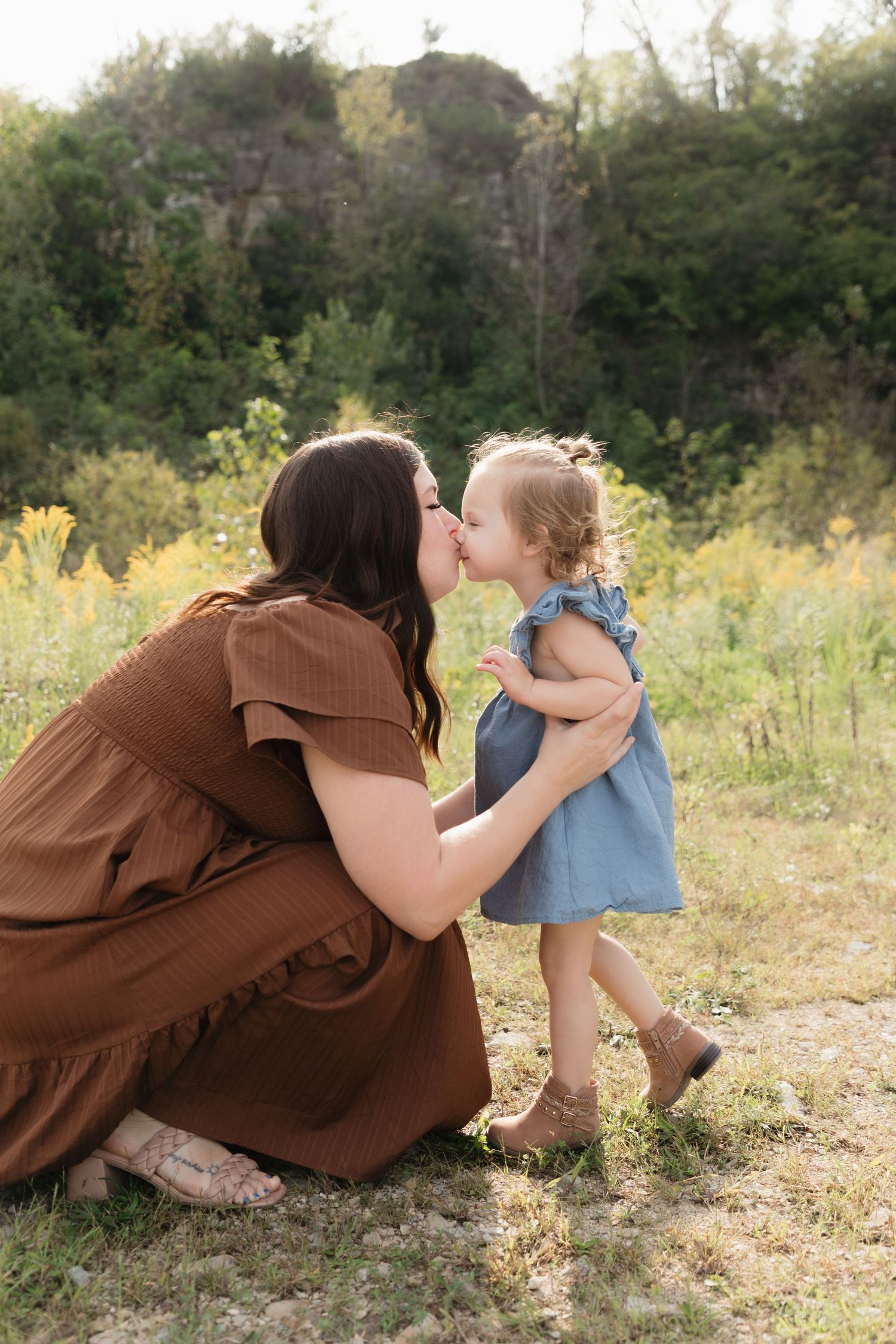 A mother in a brown dress crouches to kiss her toddler daughter in a blue dress outdoors in a sunny field.