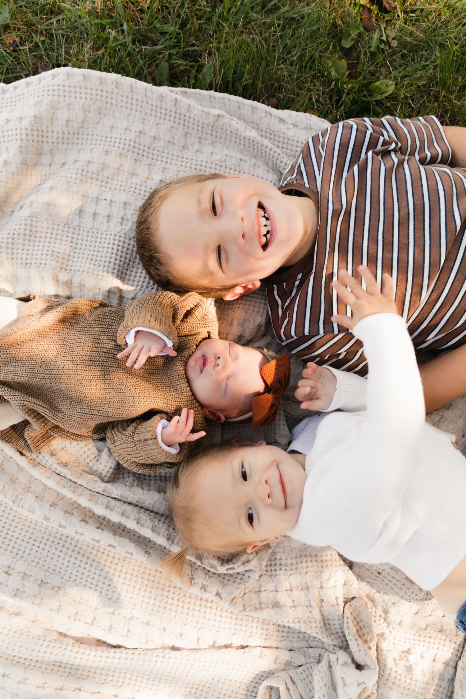 Three children lying on a blanket outdoors, two toddlers and a newborn baby, smiling together in autumn clothing.