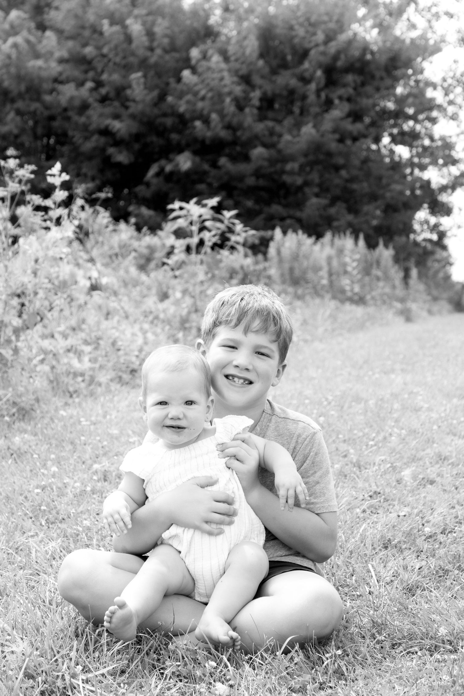 Black and white photo of a smiling boy sitting on grass holding a happy baby outdoors near trees.