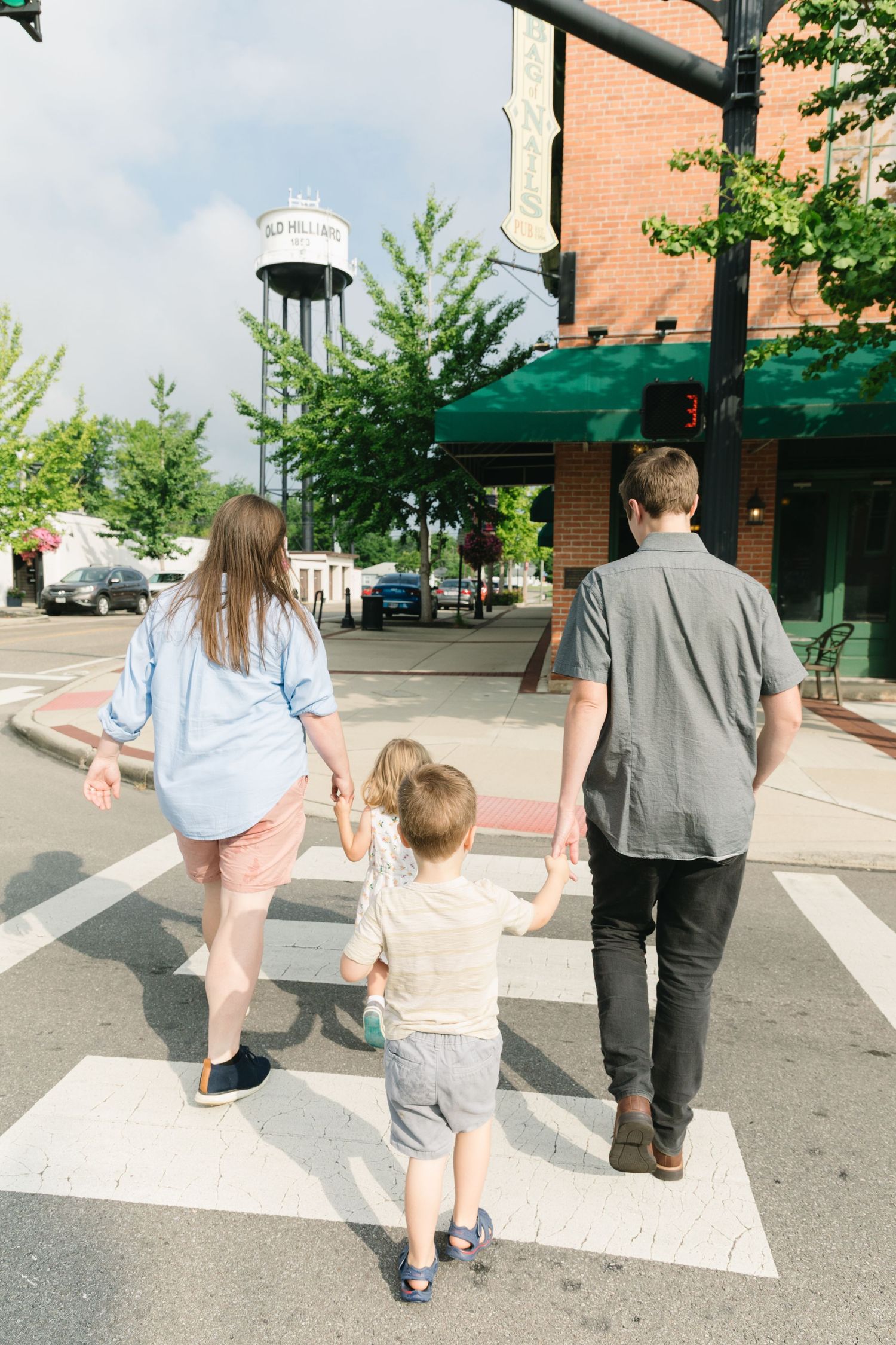 Family of three holding hands while crossing a striped crosswalk in a small town, with a water tower visible in the background.