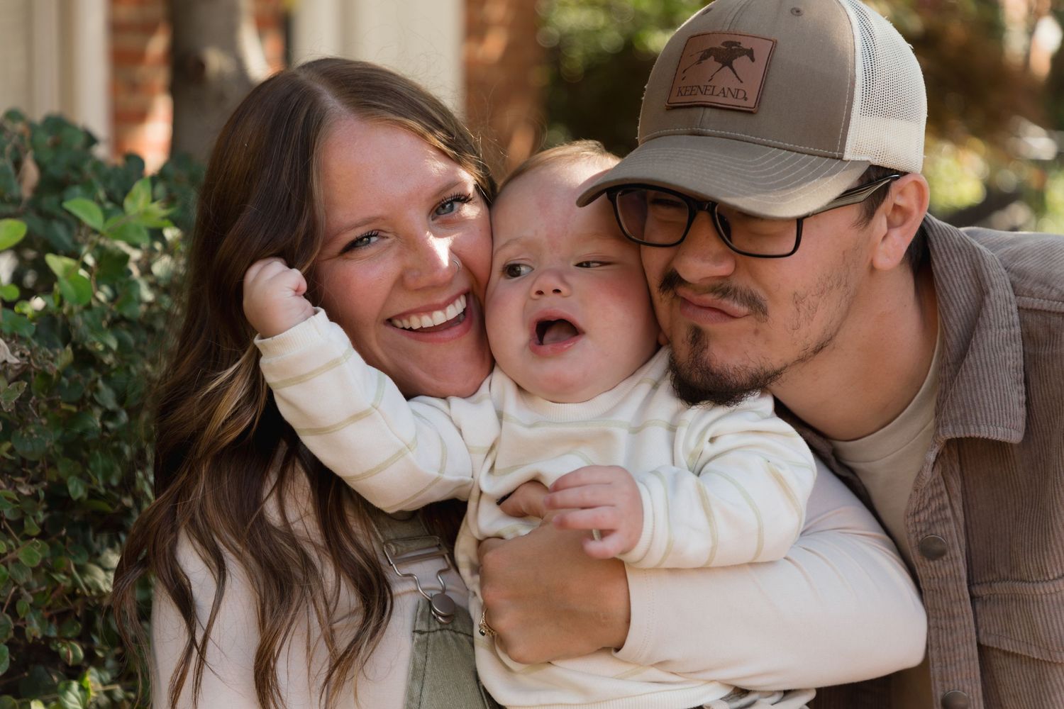 Happy young couple smiling and cuddling their laughing baby outdoors in a warm family portrait photo.