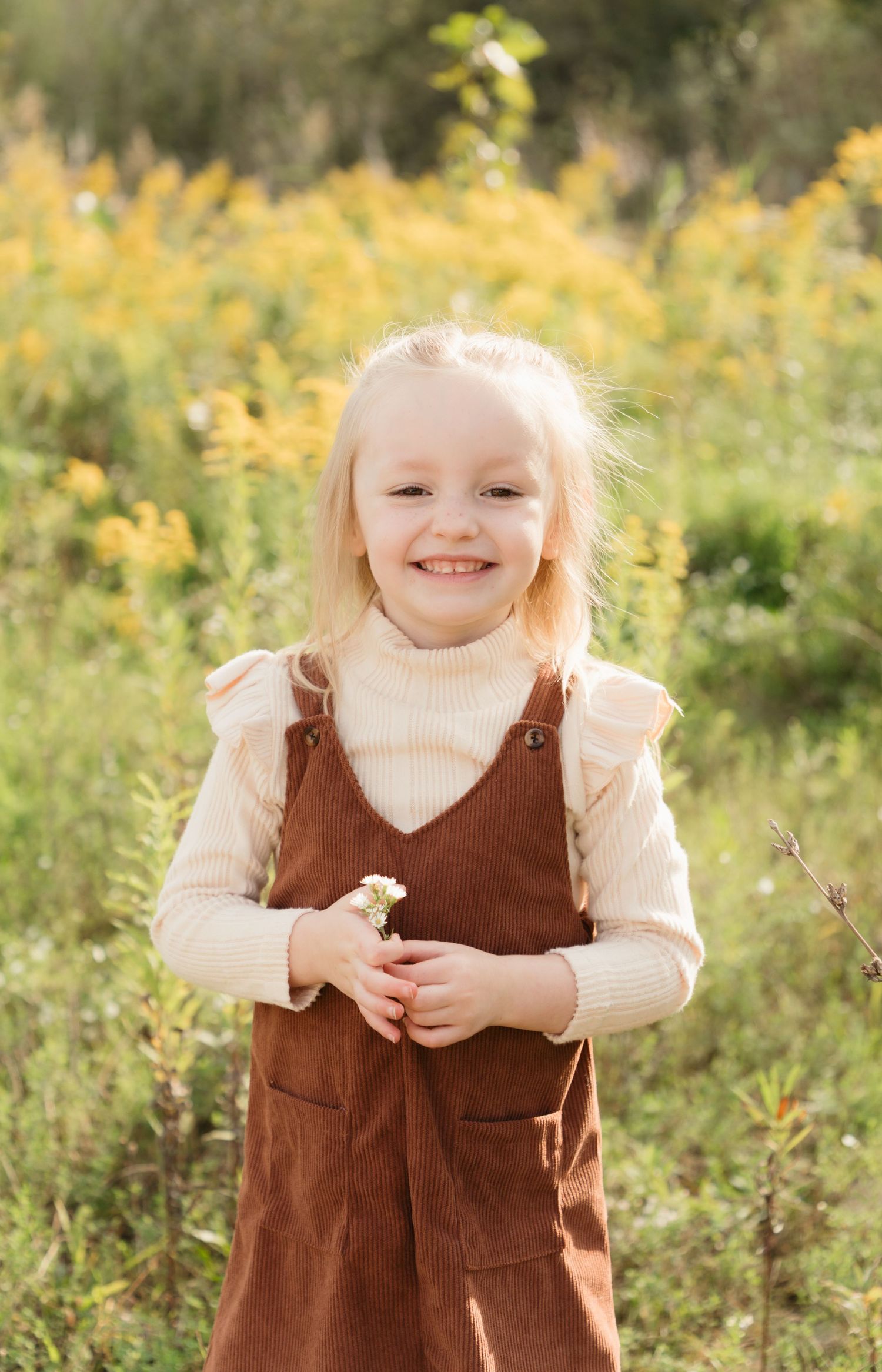 Smiling blonde girl in brown overalls and cream turtleneck holding a small flower in a sunny yellow wildflower field.