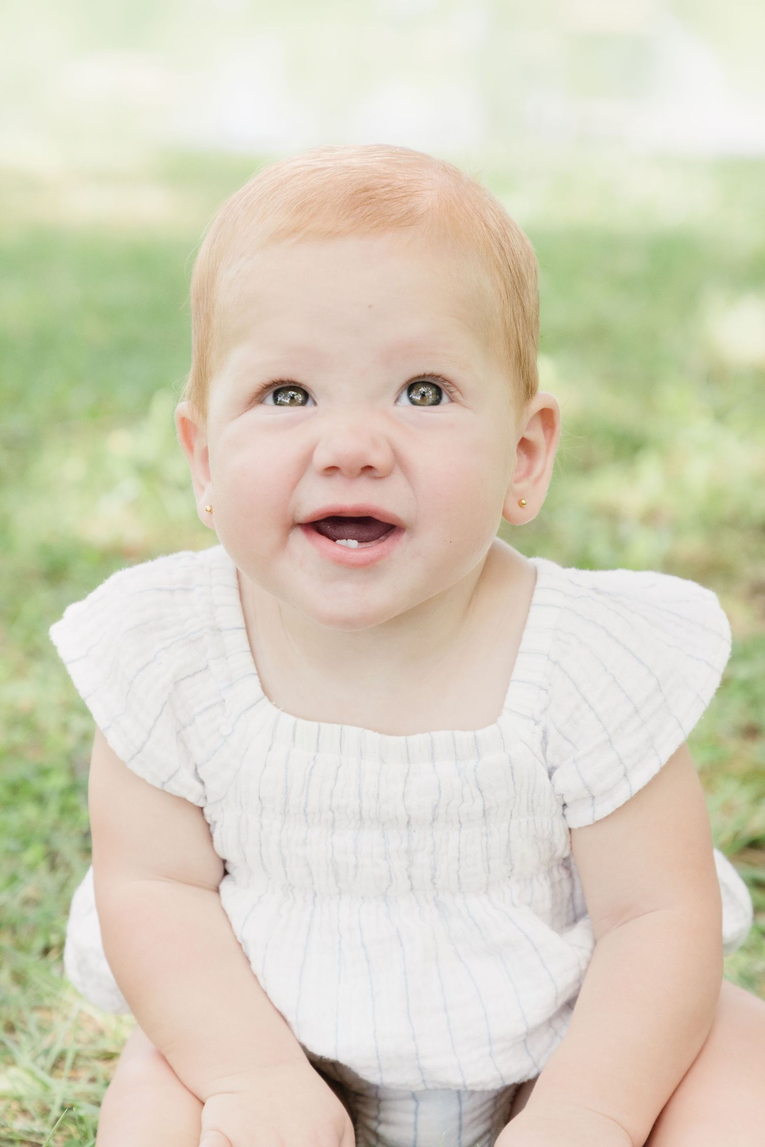Smiling baby with red hair wearing a white ruffle top, sitting on green grass outdoors.