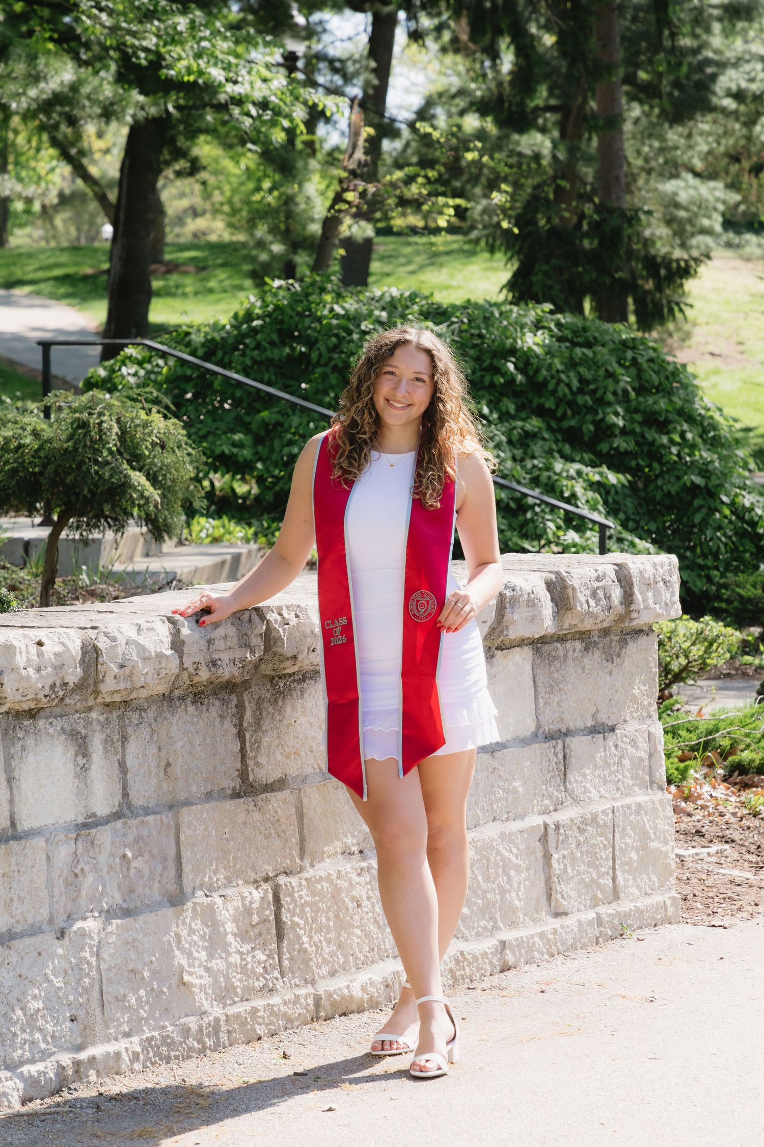 Young woman in white dress and red graduation stole smiling outdoors near stone wall with green trees.
