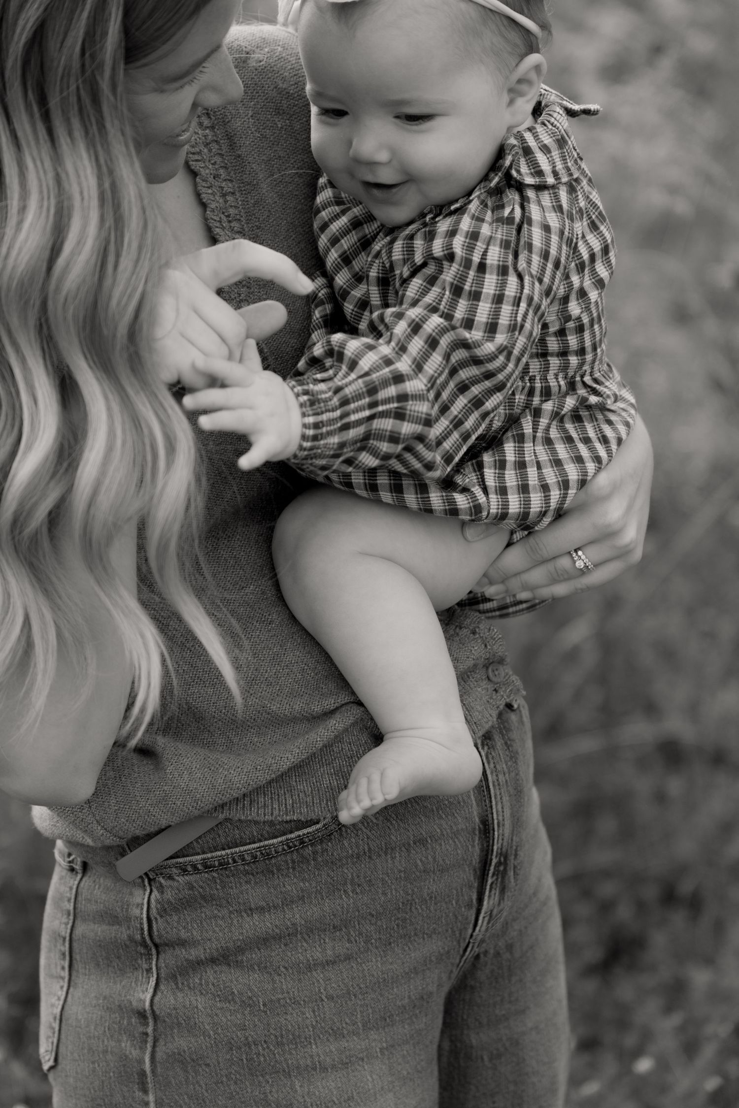 Black and white photo of a mother holding her smiling baby dressed in a plaid shirt outdoors.