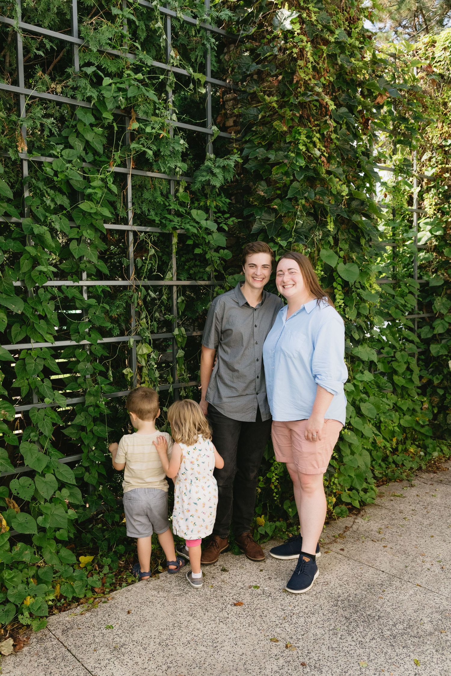 A young family poses in front of a lush green ivy-covered trellis wall, with two small children exploring the foliage.