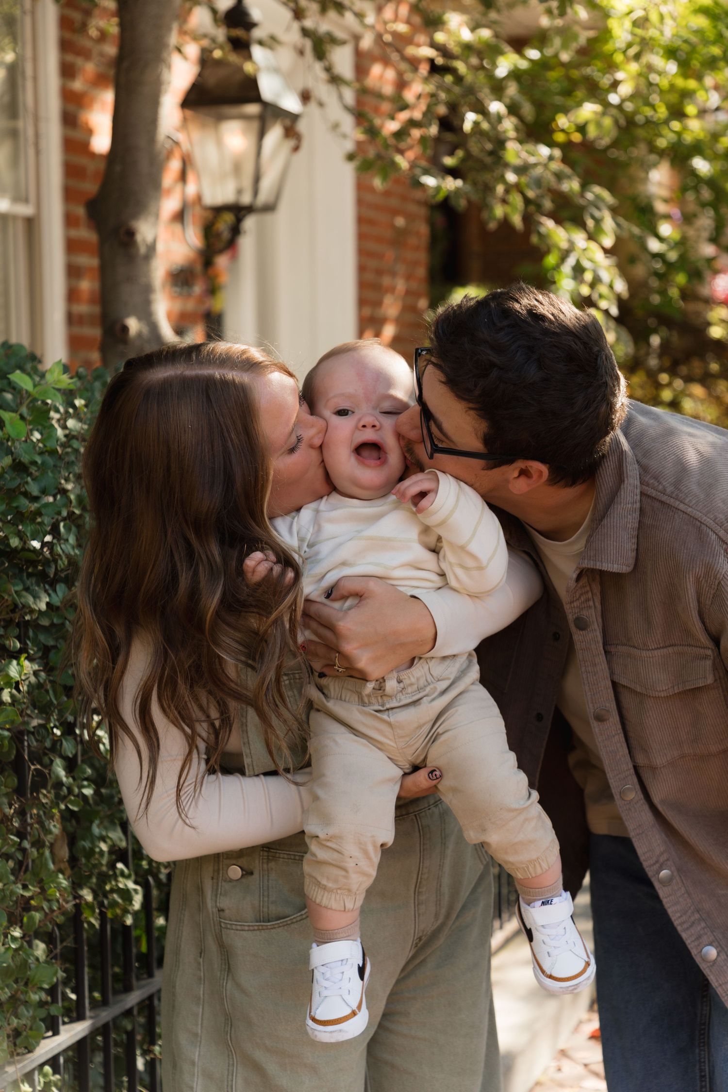 Parents kissing their laughing baby on the cheeks outdoors near brick building with green foliage.