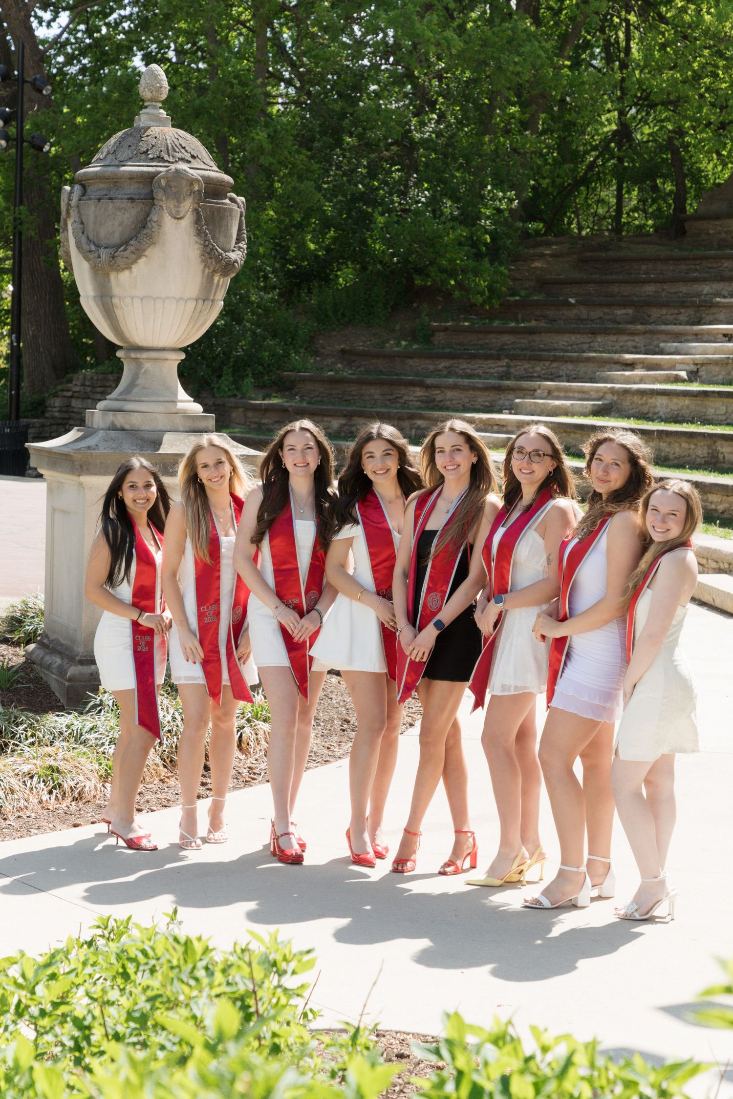 Group of young women in white outfits and red graduation sashes posing outdoors near a stone urn monument.