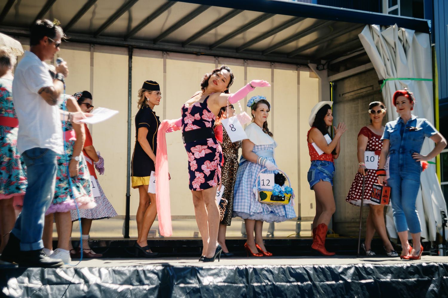Contestants in colorful retro outfits compete in a vintage costume pageant on an outdoor stage.