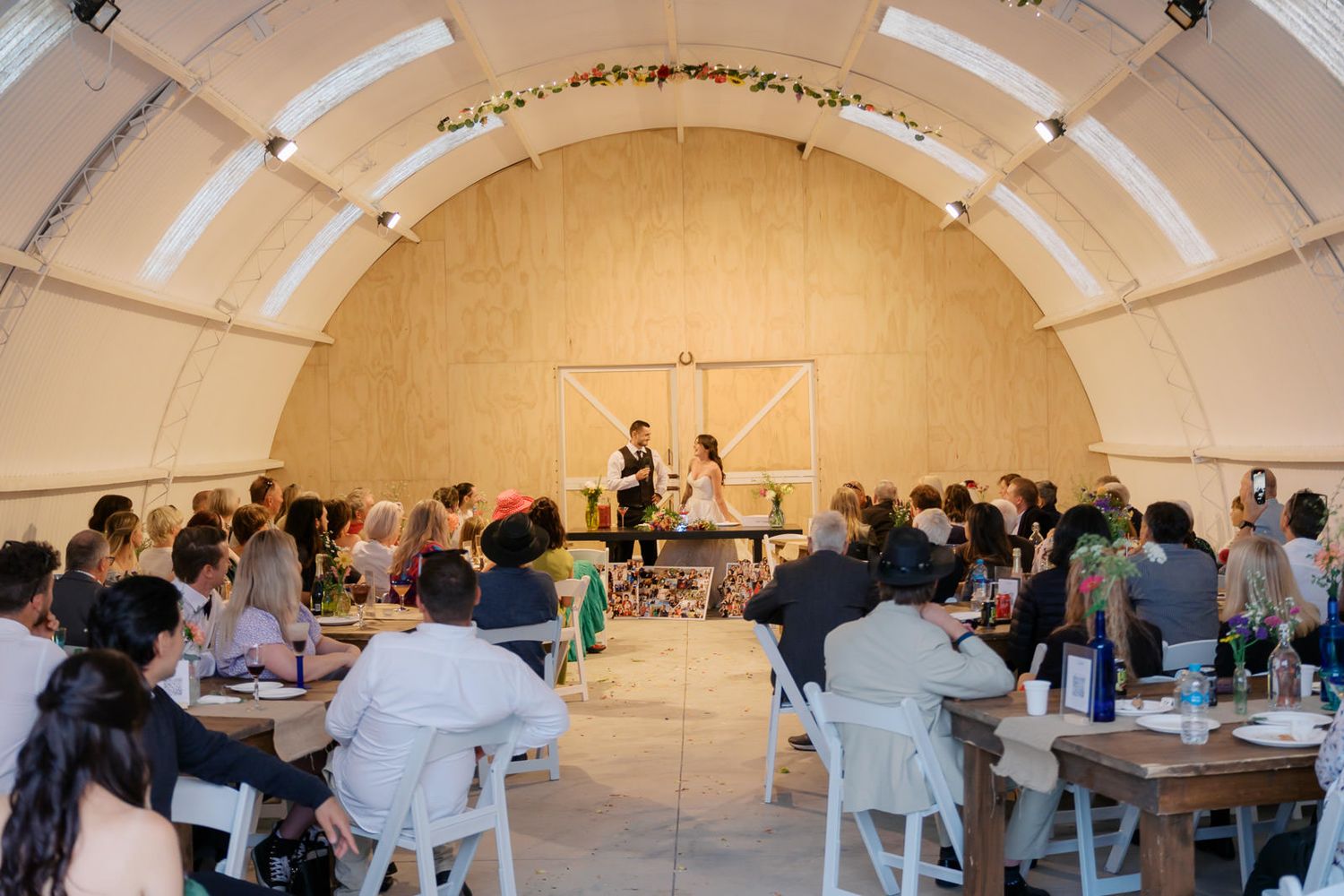 Guests gather in a rustic arch venue for an intimate wedding ceremony with floral decor and wooden tables.