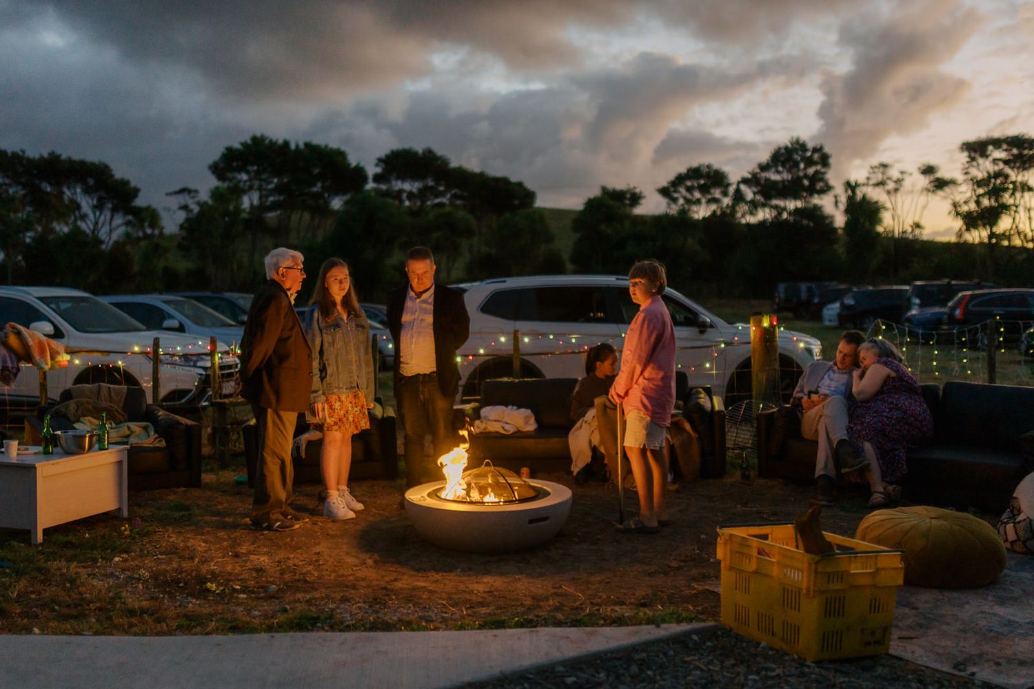 Group of people gathered around a glowing fire pit at dusk in an outdoor venue with parked cars in background.