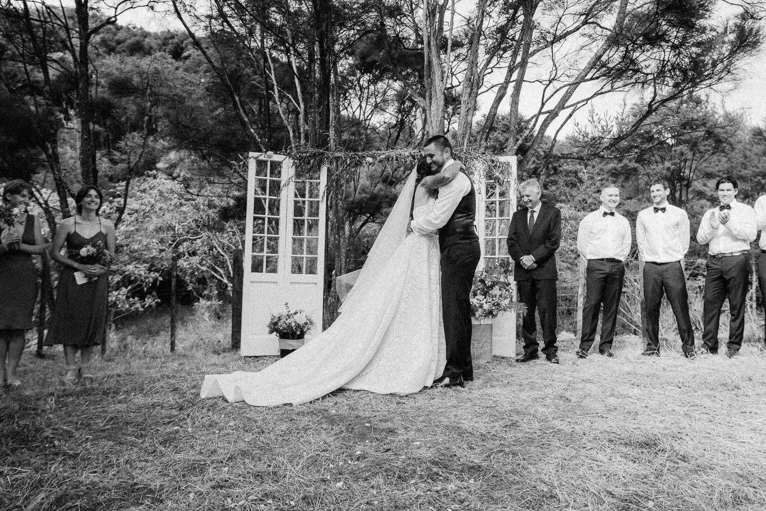 Black and white outdoor wedding ceremony with bride and groom kissing, bridal party standing alongside, trees in background.